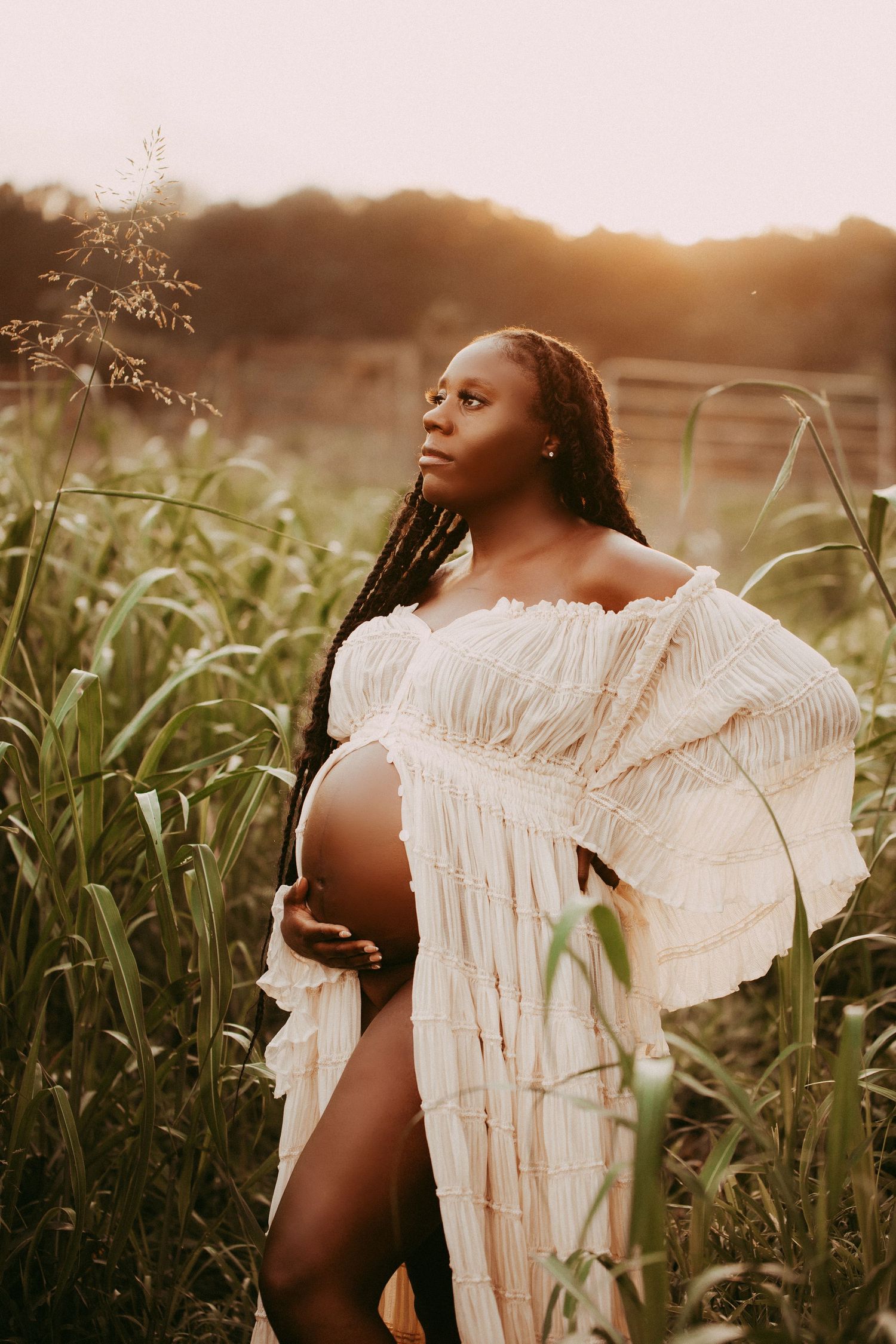 A woman in a white ruffled dress poses in a golden-lit field at sunset for an intimate maternity photoshoot.