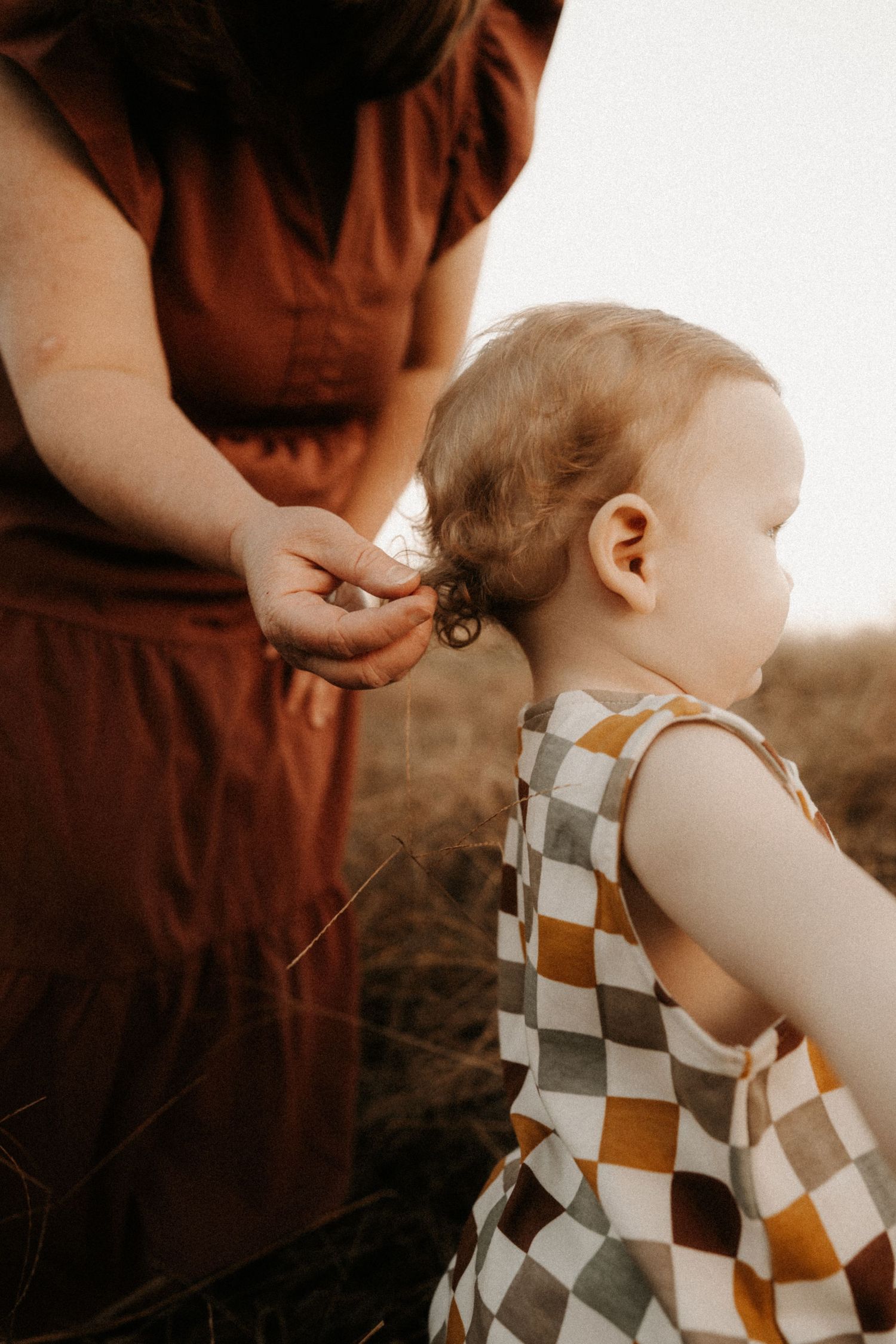 A parent in a brown dress adjusts the ear of a small child wearing a checkered pattern romper in a sepia-toned setting.