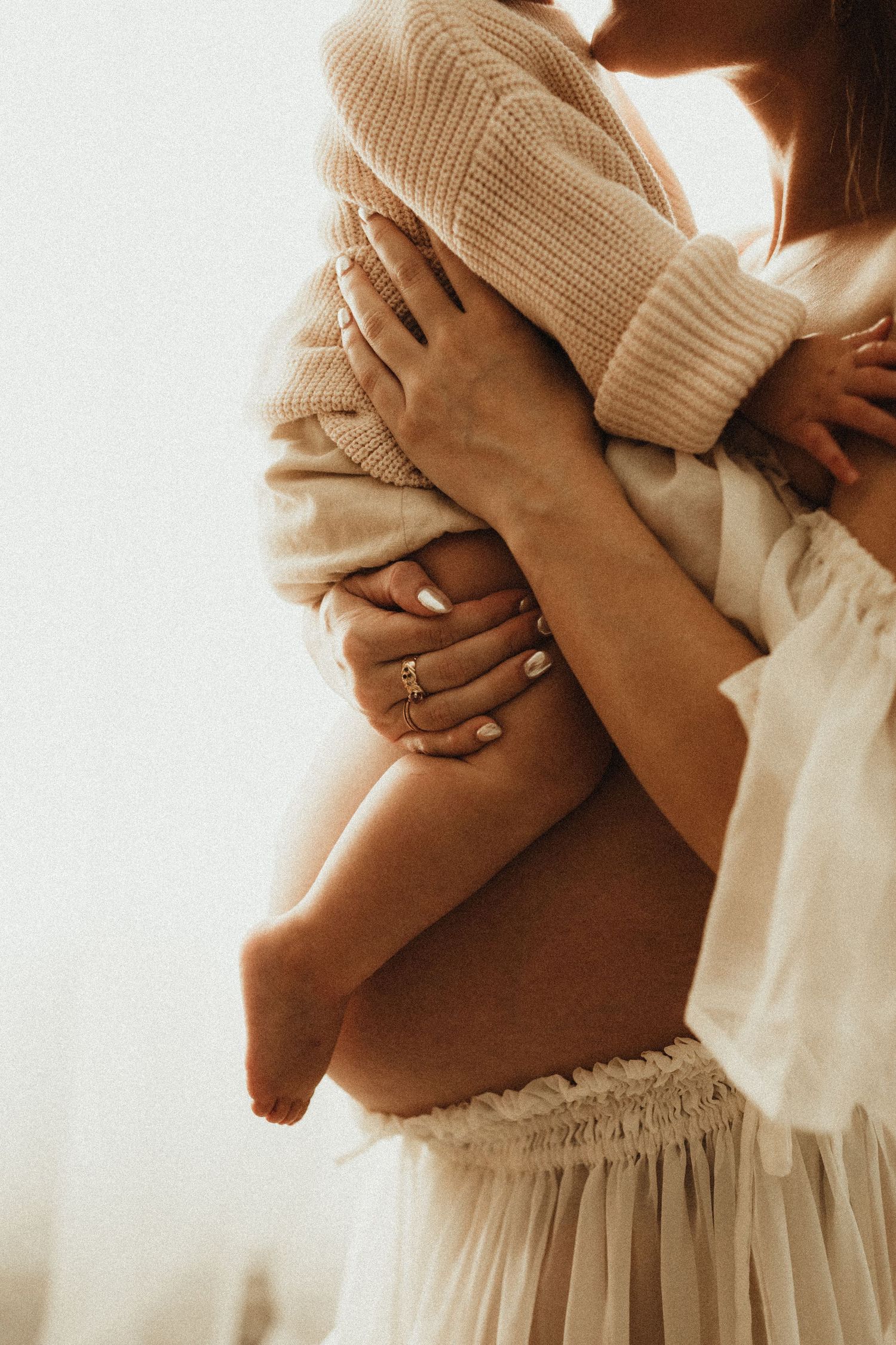 Close-up of person wearing beige sweater and white skirt hugging themselves in soft, warm lighting.