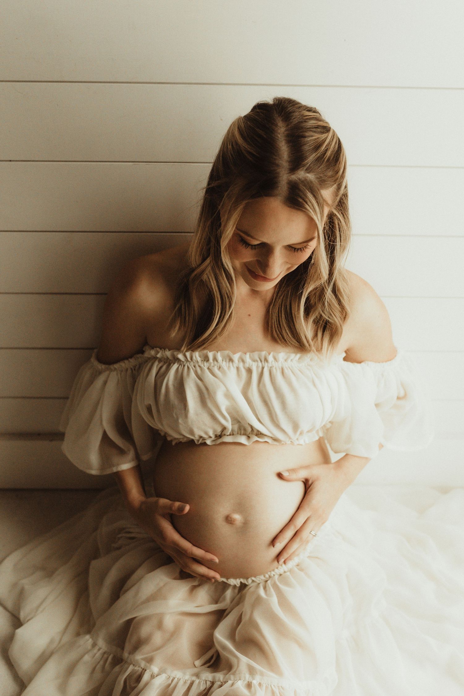 Artistic maternity photo series showing a pregnant woman in white dress against shiplap wall captured in muted tones.