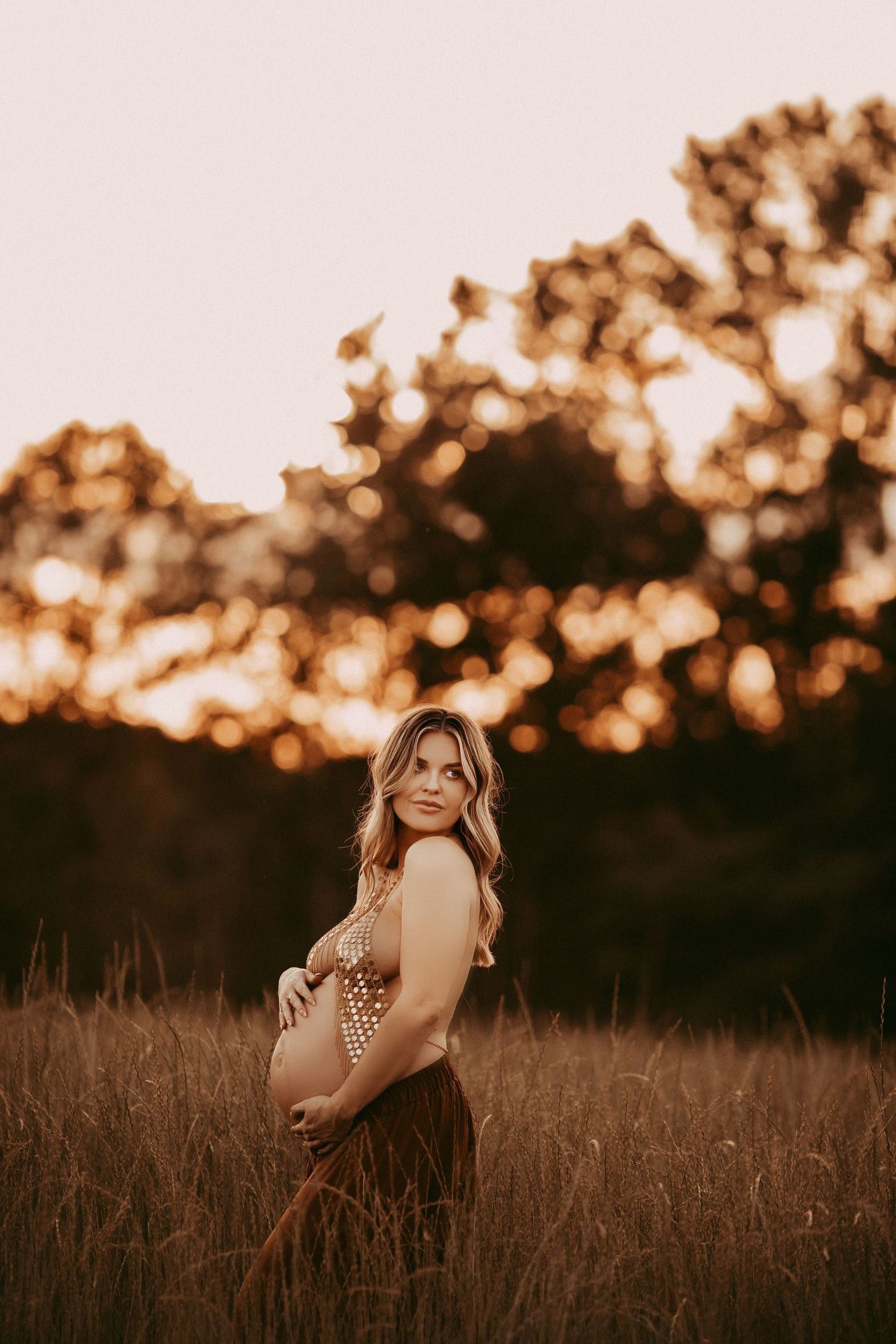 Woman in floral dress poses in golden field at sunset with dramatic backlighting and warm autumn colors.