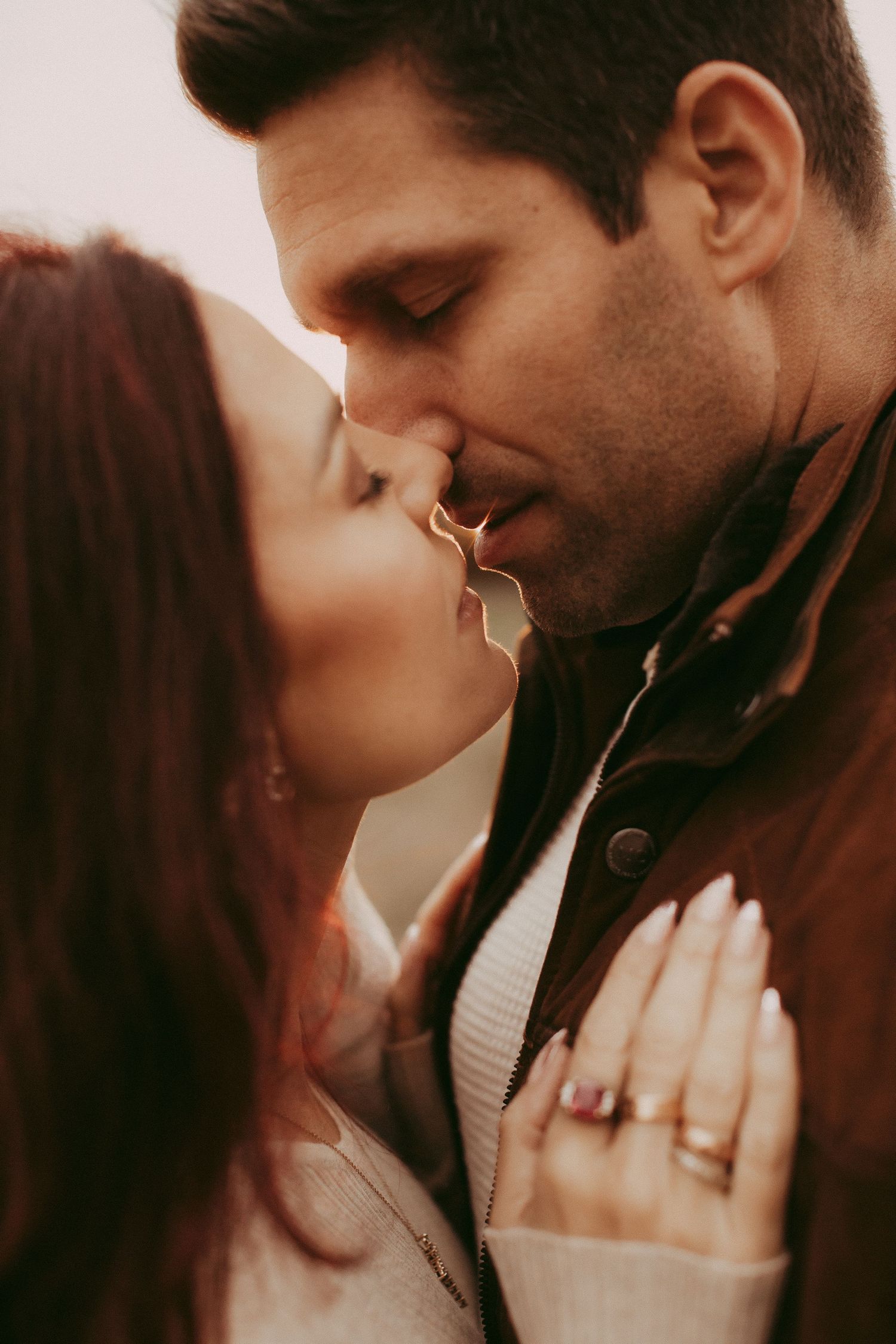 A romantic couple sharing a tender kiss, woman wearing an elegant engagement ring and manicured nails, warm toned intimate photo.
