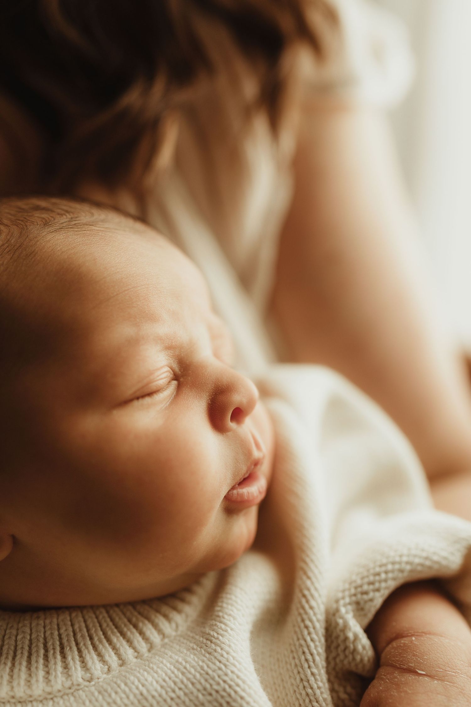 Sleeping newborn baby in cream knit outfit cradled in mother's arms in soft warm light.