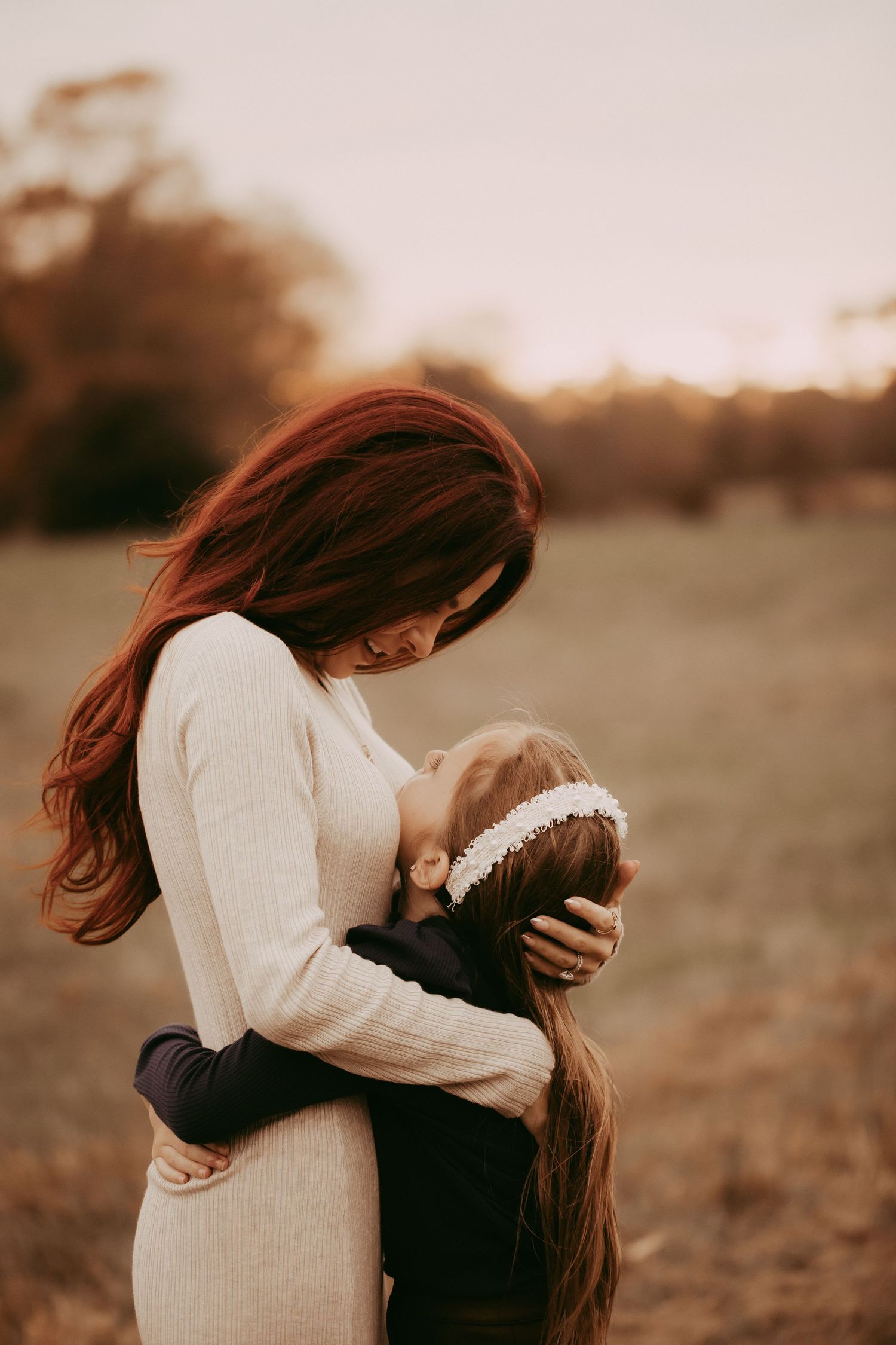 A mother with red hair in a white dress tenderly embraces her young daughter outdoors at golden hour.