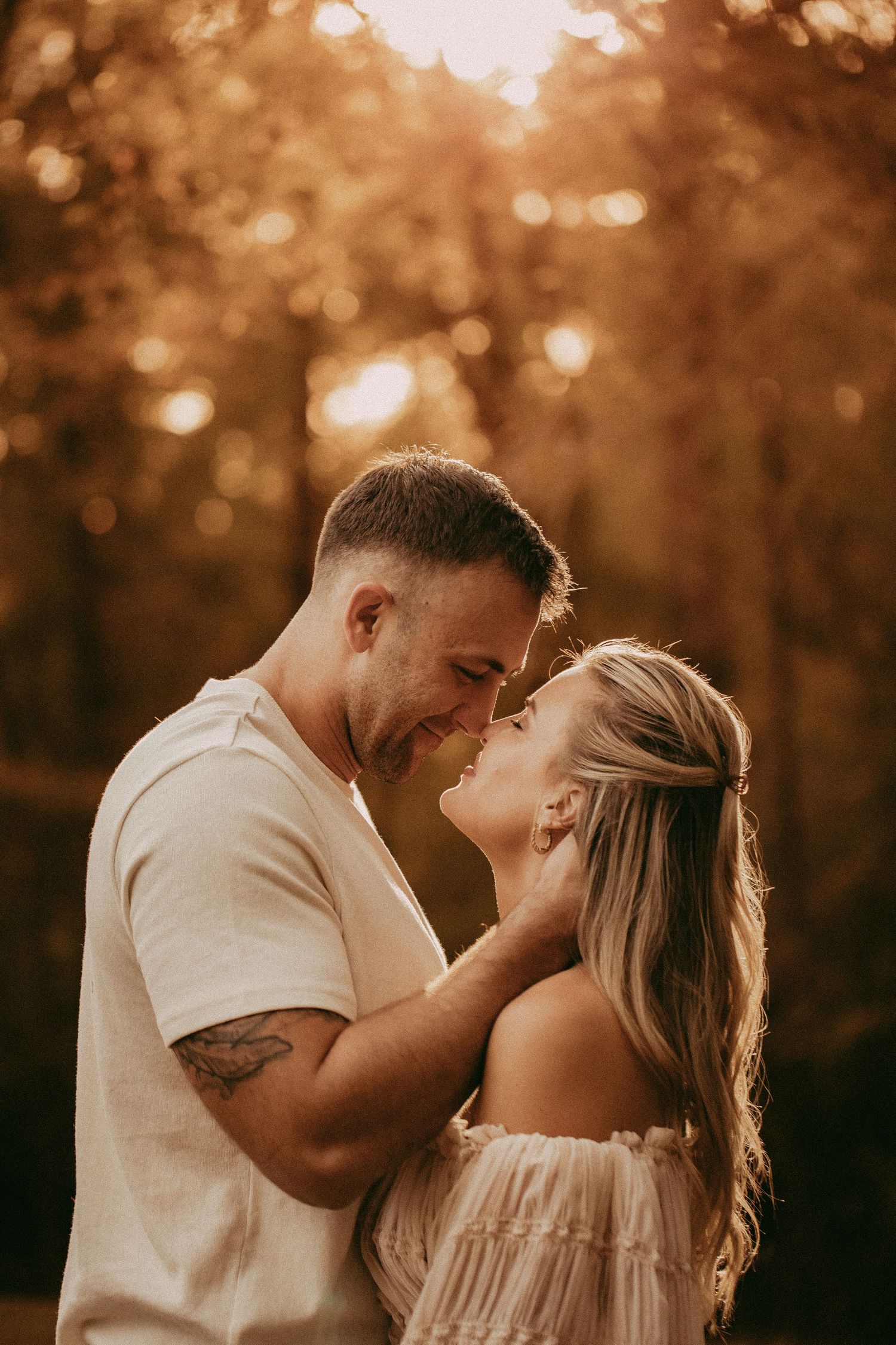 A couple shares a romantic forehead-to-forehead moment outdoors, surrounded by warm golden autumn bokeh lighting.