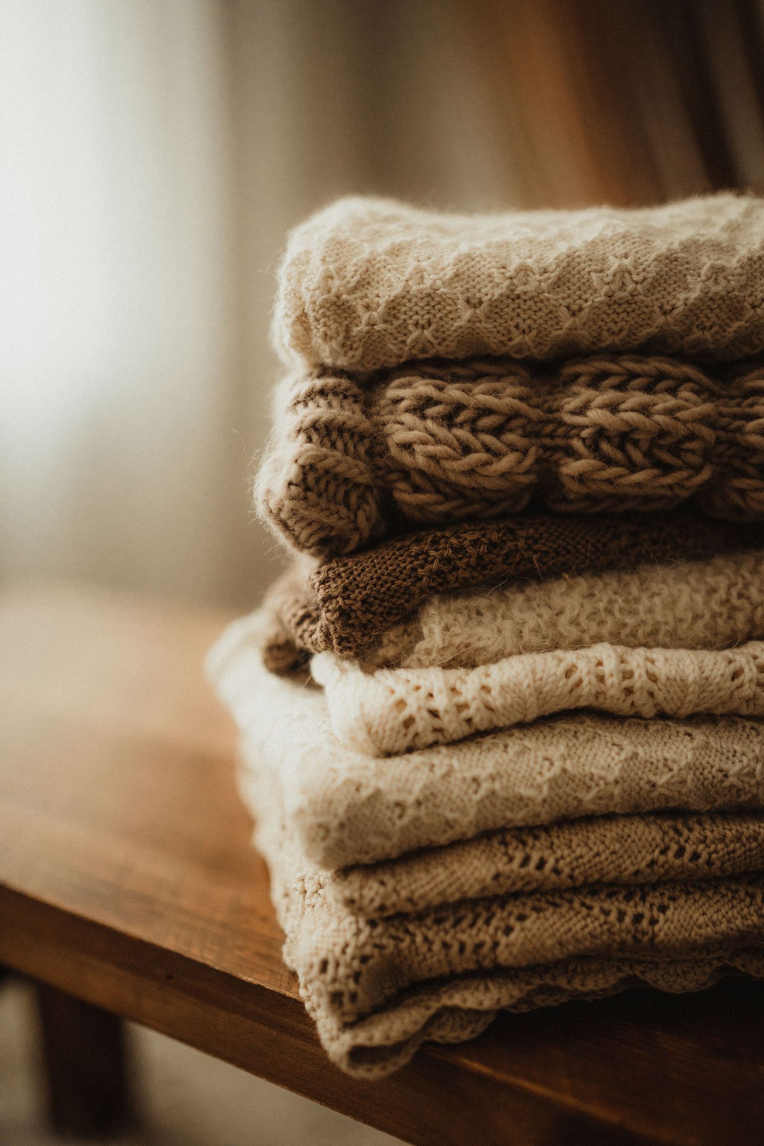 A stack of cozy knitted beige sweaters folded neatly on a wooden surface in warm lighting.