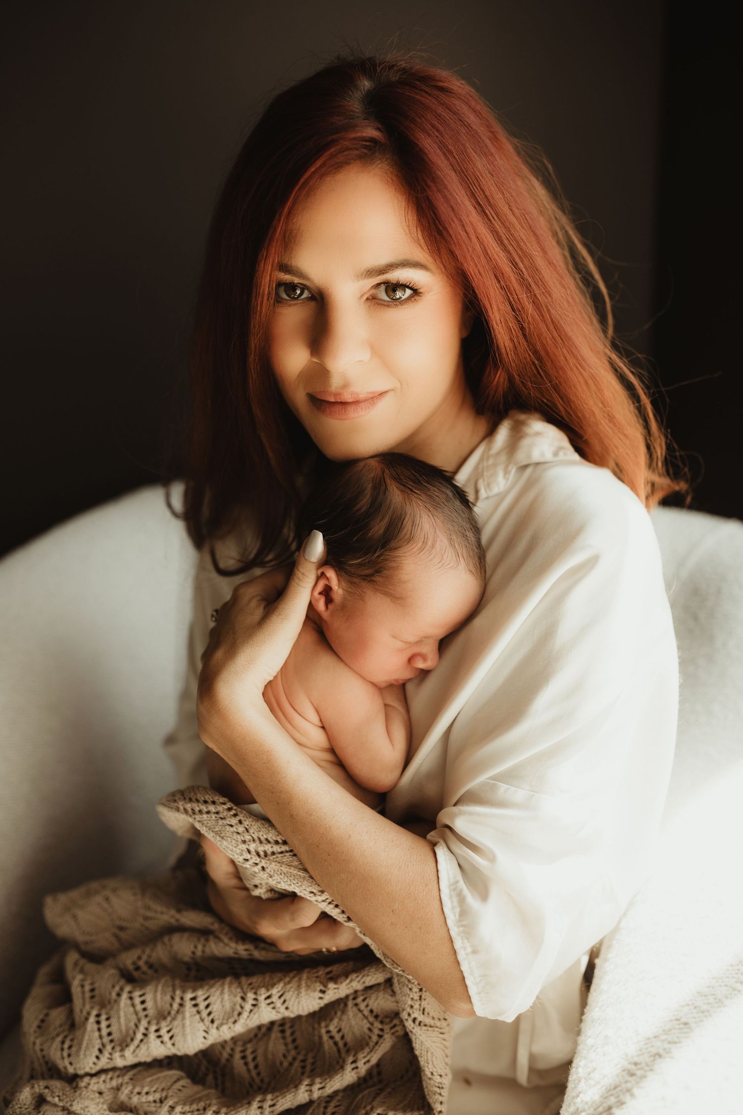 A mother in a white shirt holds sleeping newborn baby against soft knit blankets in warm dramatic lighting.