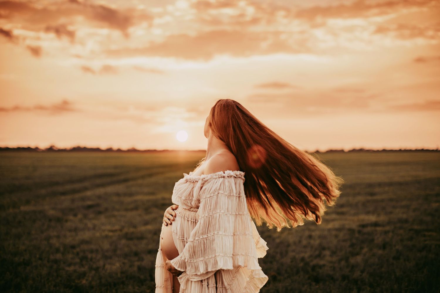 Long flowing hair catches the wind as figure in white dress stands in field against dramatic orange sunset sky.