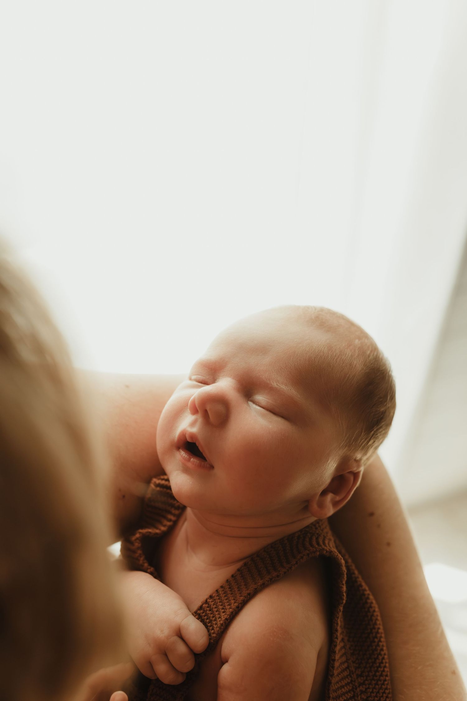 A newborn baby sleeping peacefully in a parent's arms, wearing a brown knit outfit in soft natural light.
