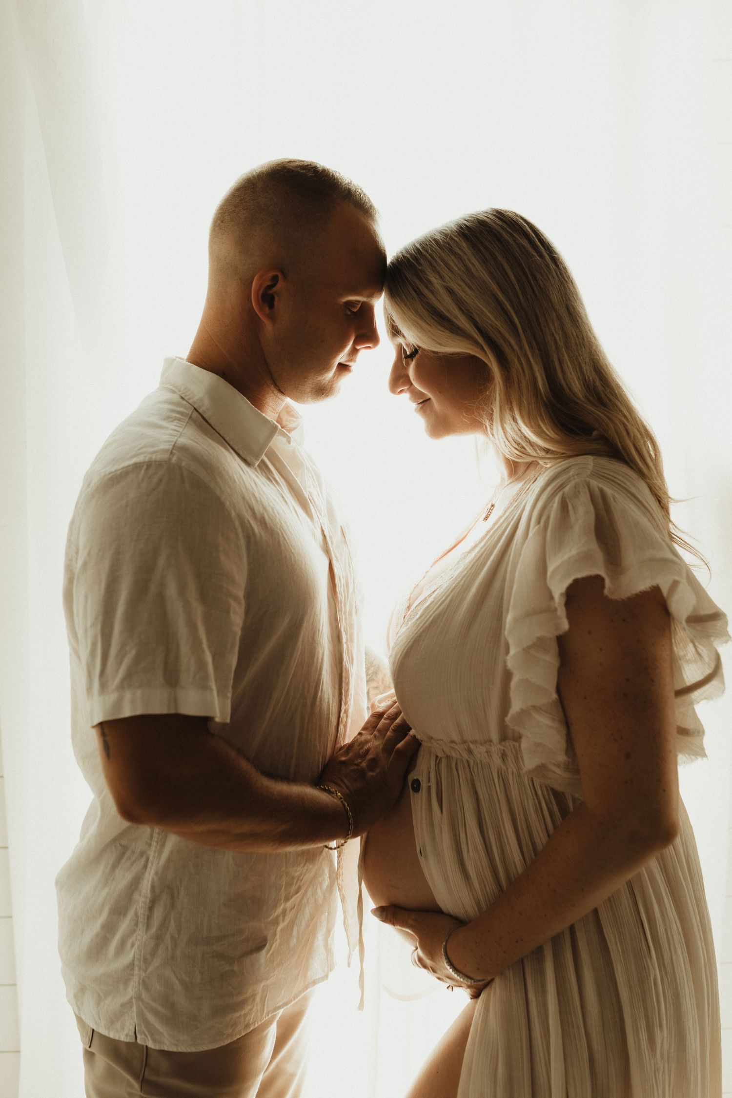 A couple in white outfits share a tender moment, foreheads touching, as the woman cradles her pregnant belly near a bright window.