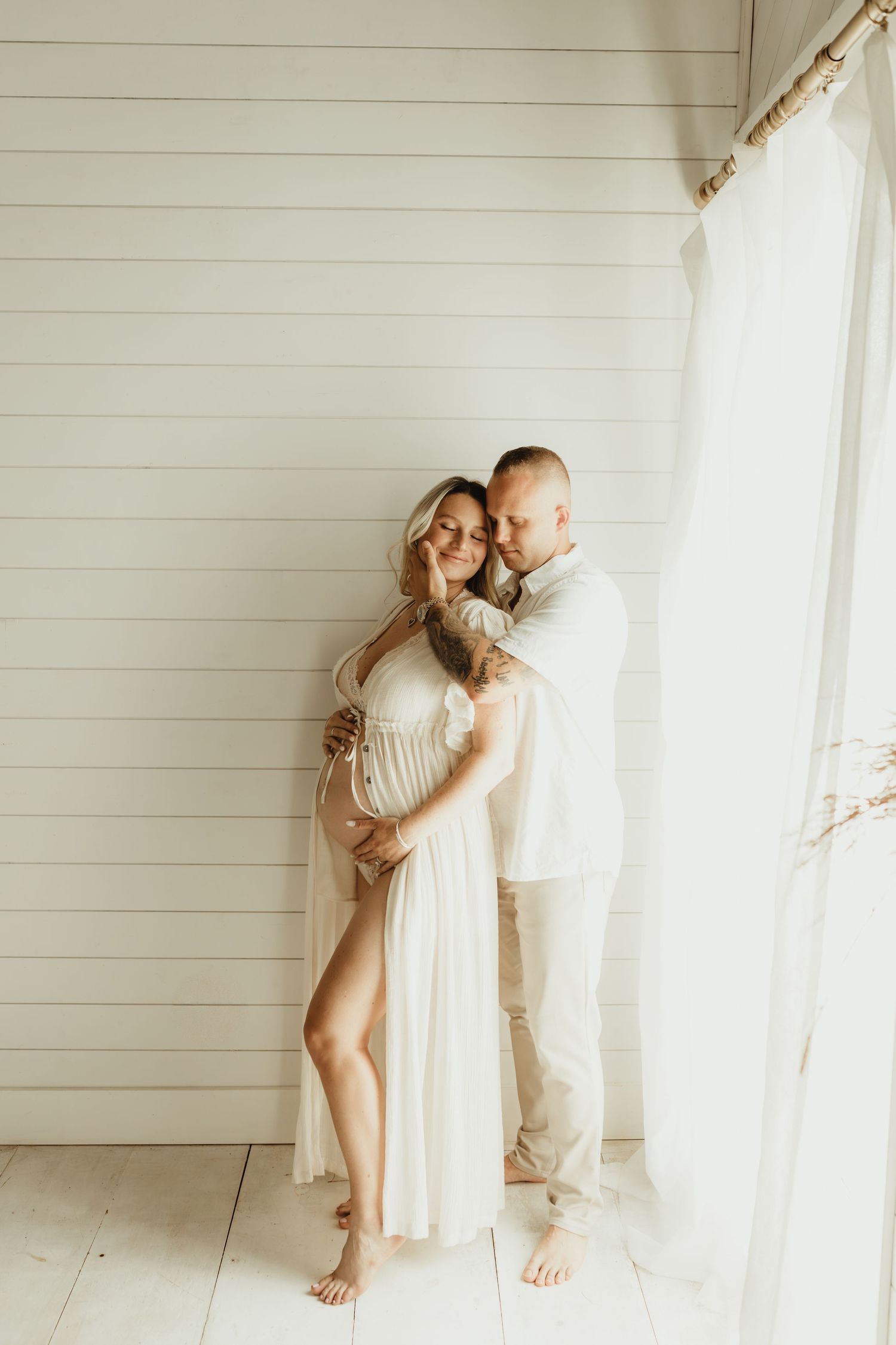Couple embracing tenderly in bright white room, pregnant woman in flowing white gown, partner holding her from behind near sheer curtains.