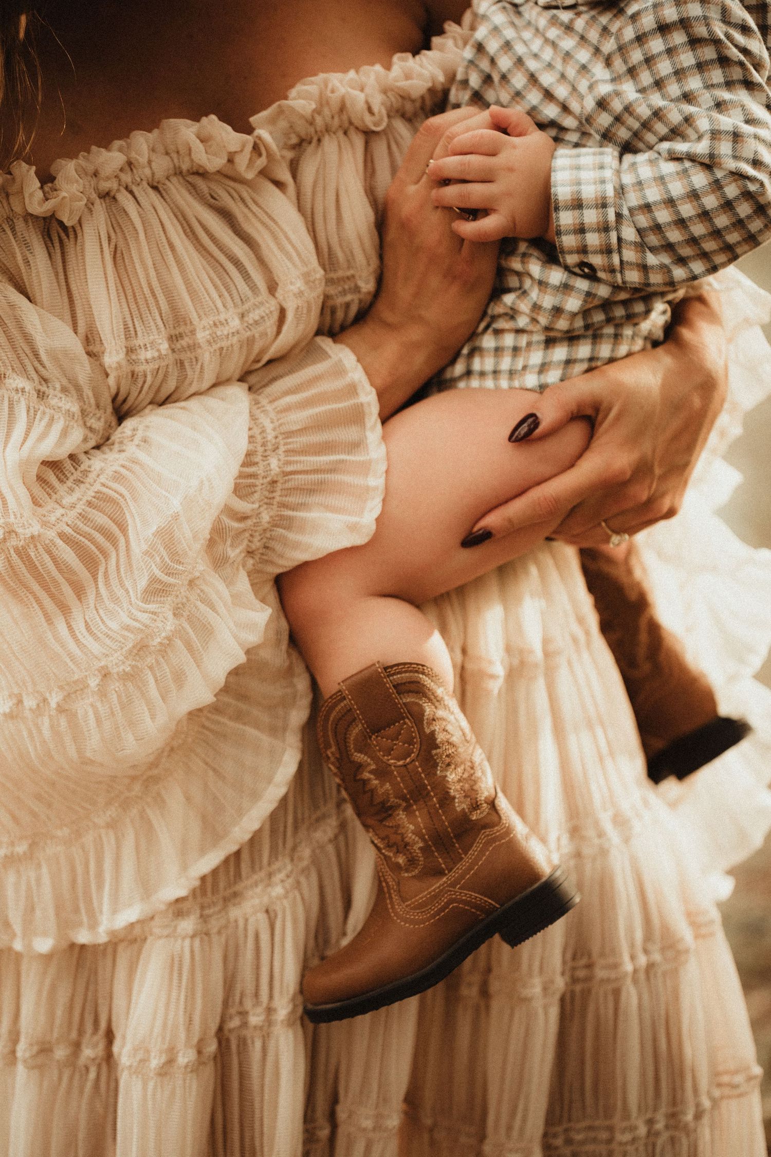 Woman in white ruffled dress holds baby wearing brown cowboy boots, showcasing a charming western-inspired family moment.