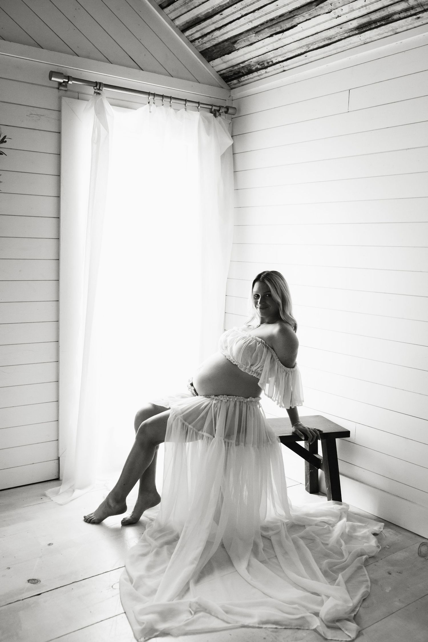 Black and white maternity photo of pregnant woman in white flowing dress sitting on bench near bright window.