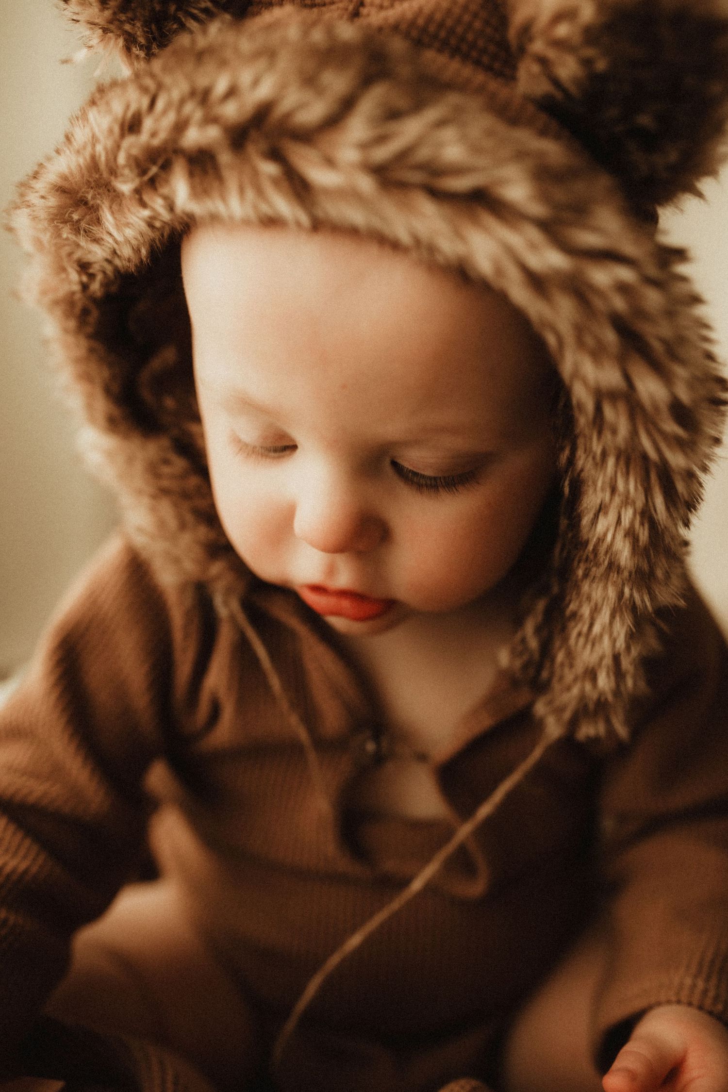 A small child wearing a cozy brown hooded sweater with fur trim looks down in a warm-toned portrait series.