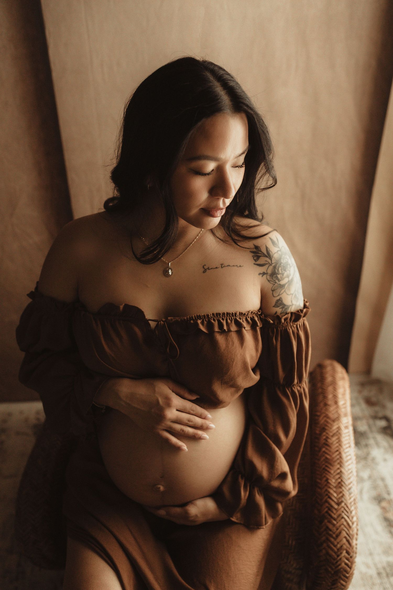Pregnant woman in brown off-shoulder dress cradling her baby bump, with tattoos and a necklace, in warm studio light.