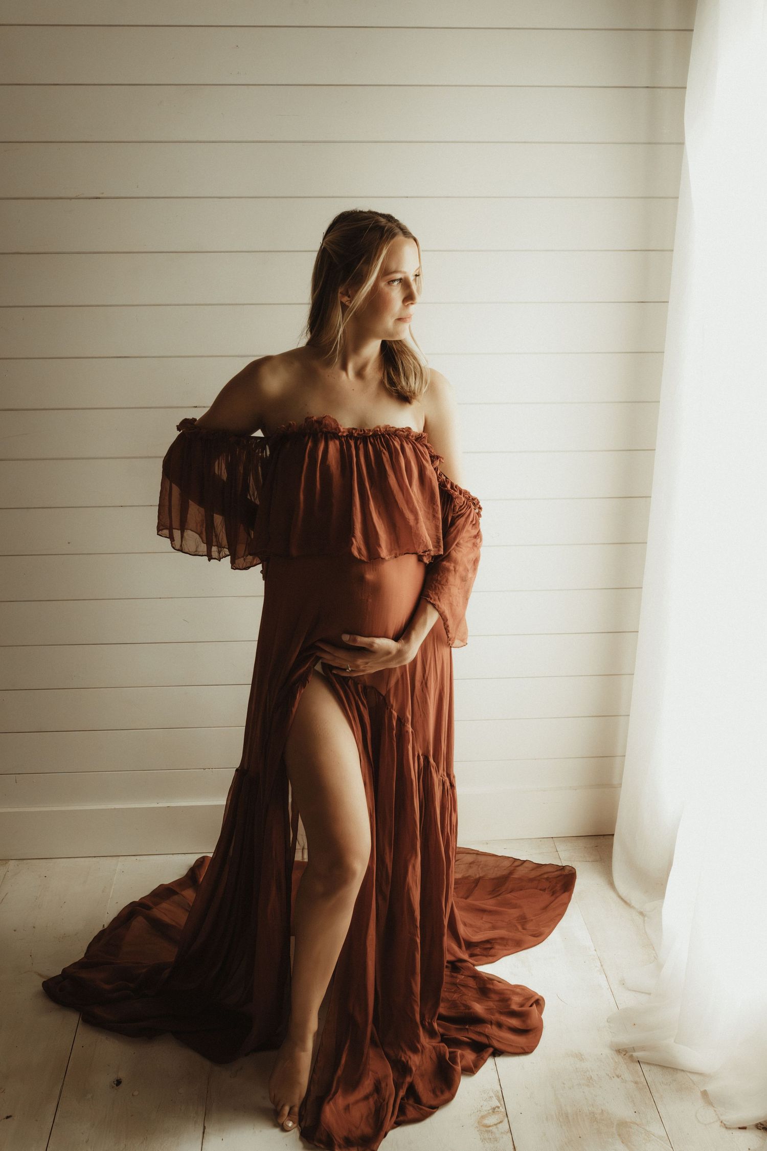 Figure in a flowing rust-colored dress poses gracefully against a white wall and wooden floor in artistic portraits.