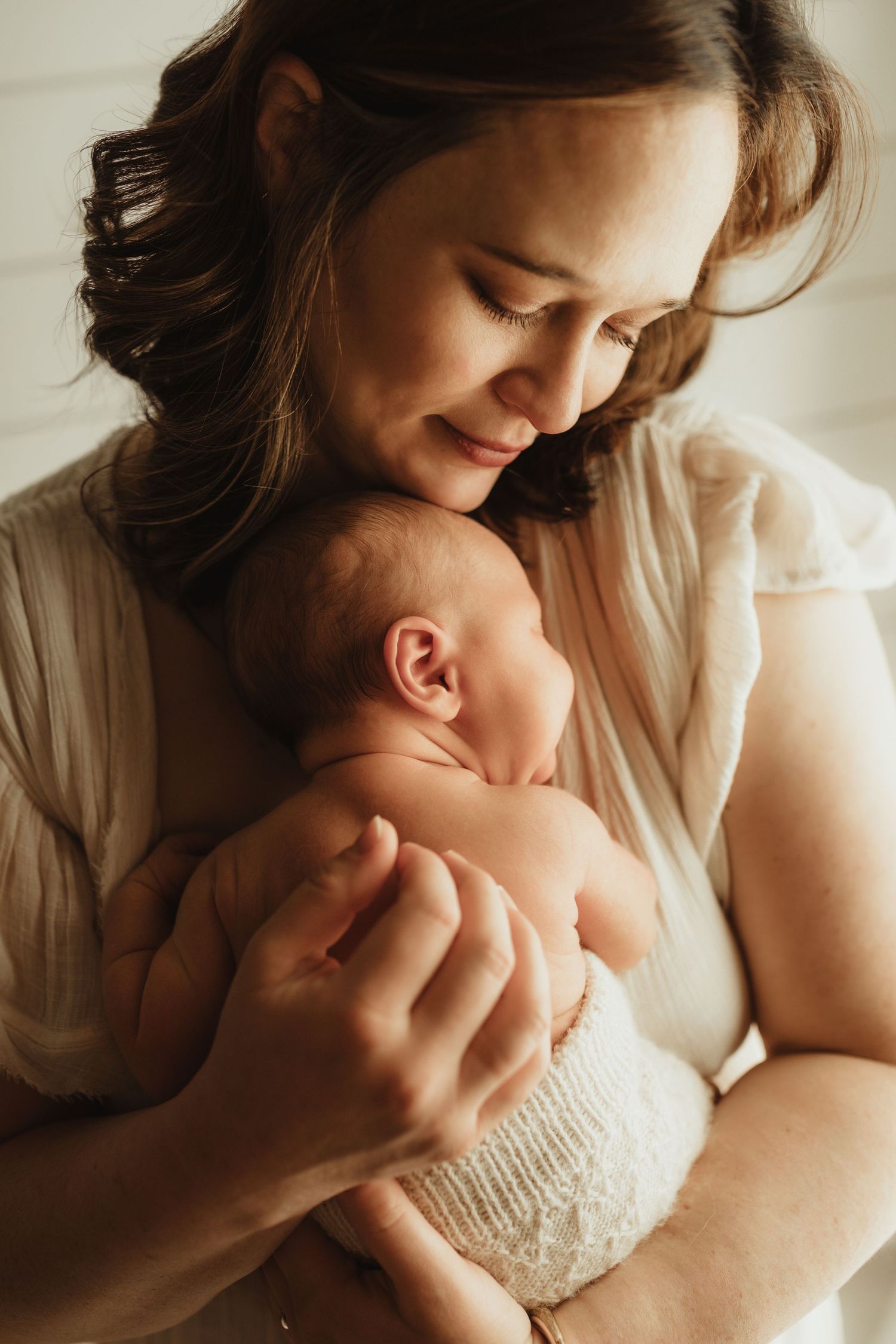 A smiling mother tenderly holds her sleeping newborn baby close to her face in a warm, softly lit portrait.