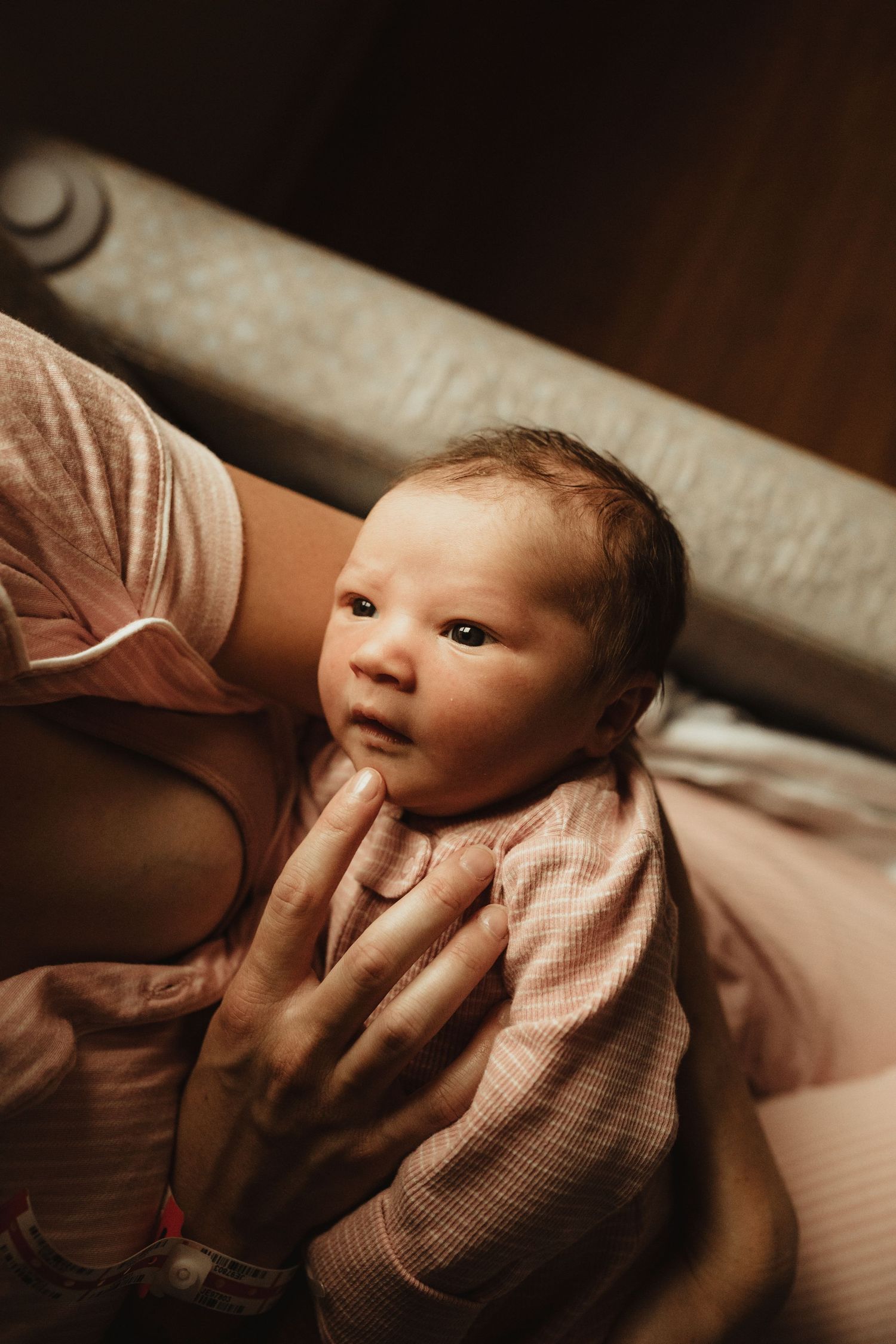 A newborn baby in pink clothing cradled gently in a parent's arms, gazing upward in warm, soft light.