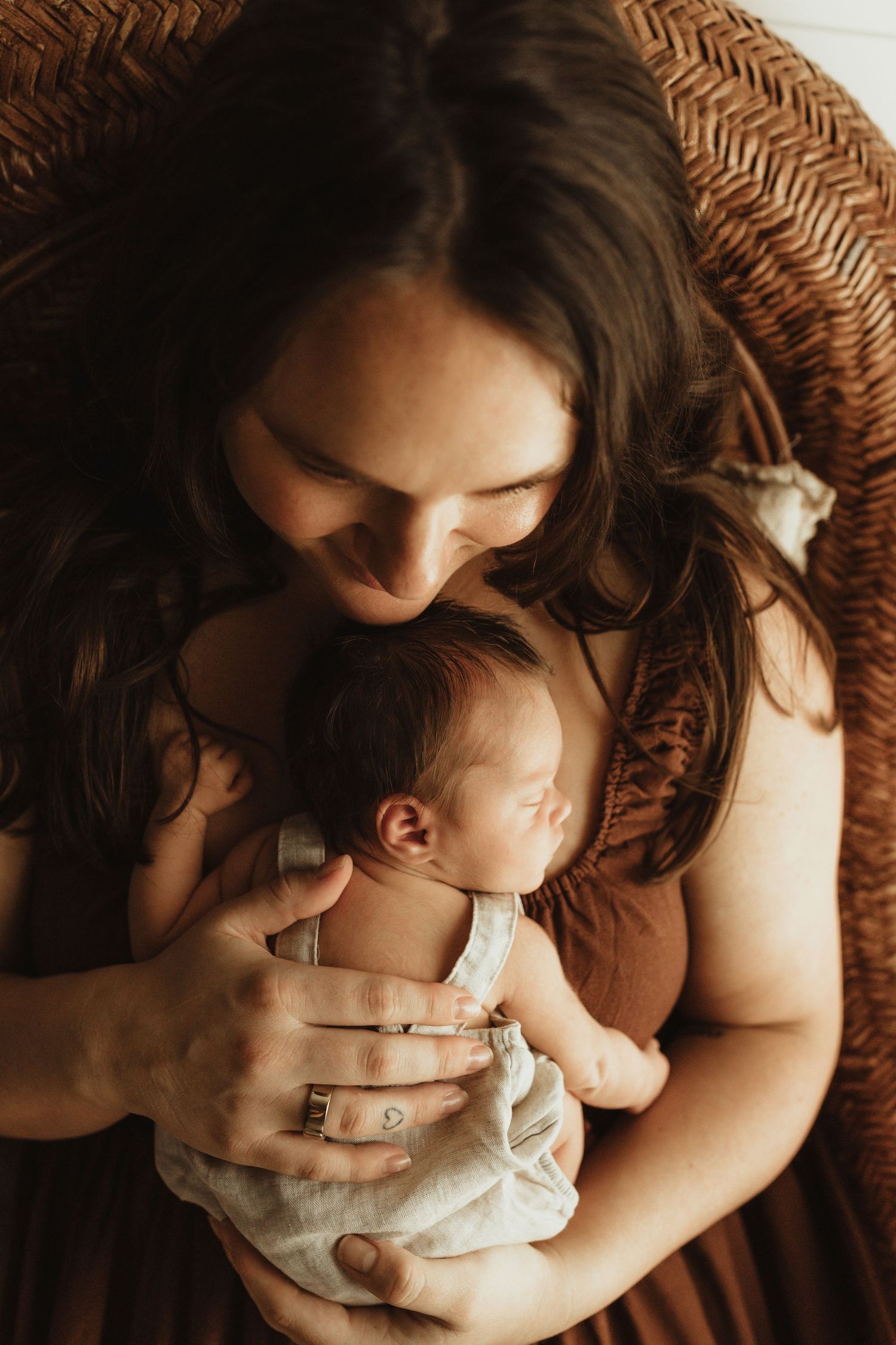 A mother tenderly holds her sleeping newborn baby in a wicker chair, wearing a rust-colored dress.