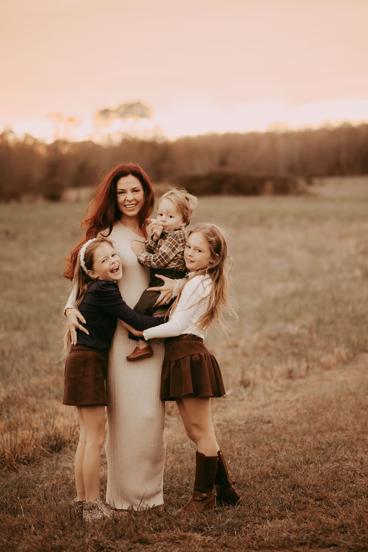 A smiling mother with red hair holds three young children in a golden field at sunset.