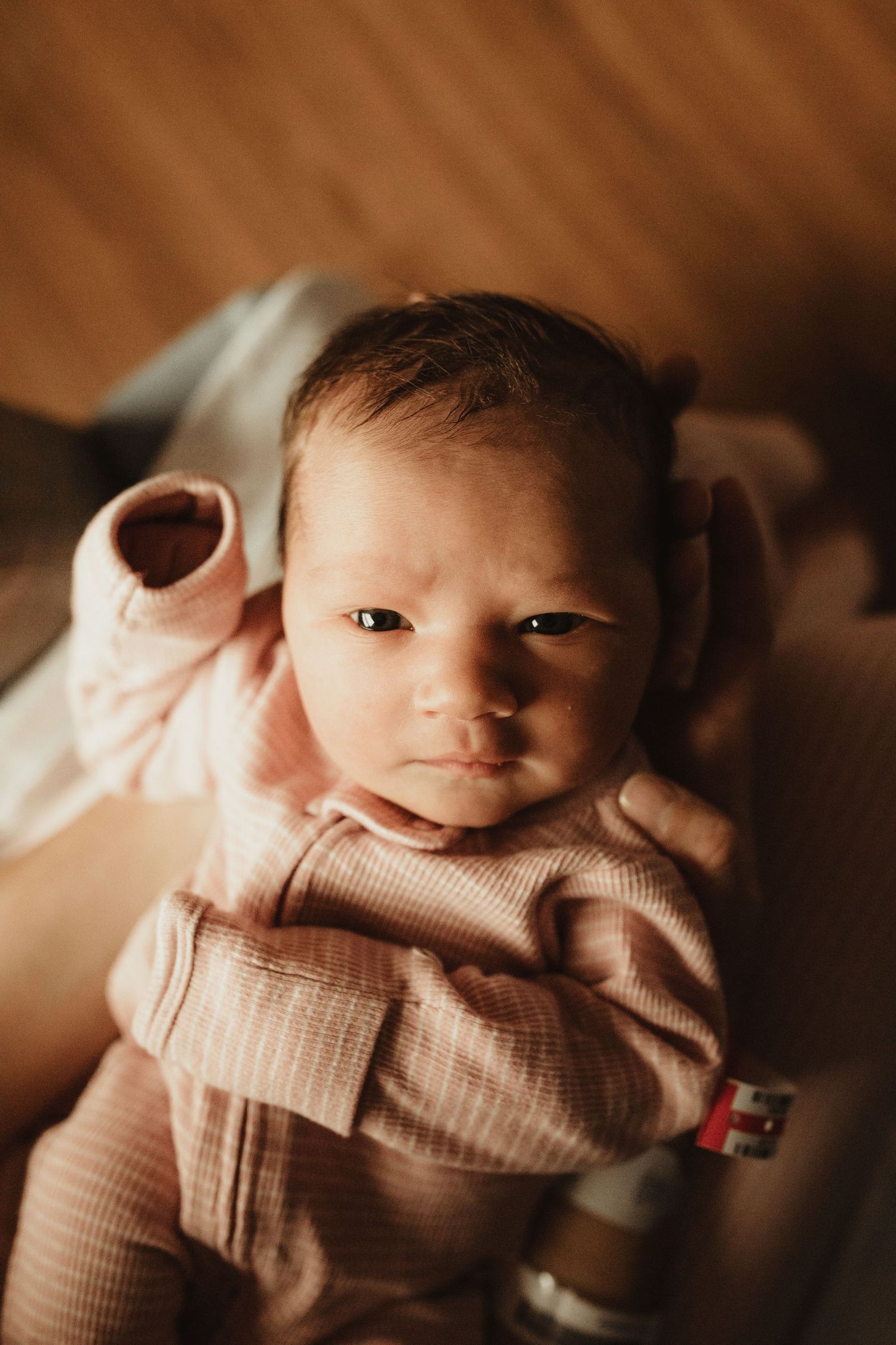 A newborn baby in a pink ribbed outfit lies on their back, gazing upward with dark eyes and tiny fists raised.