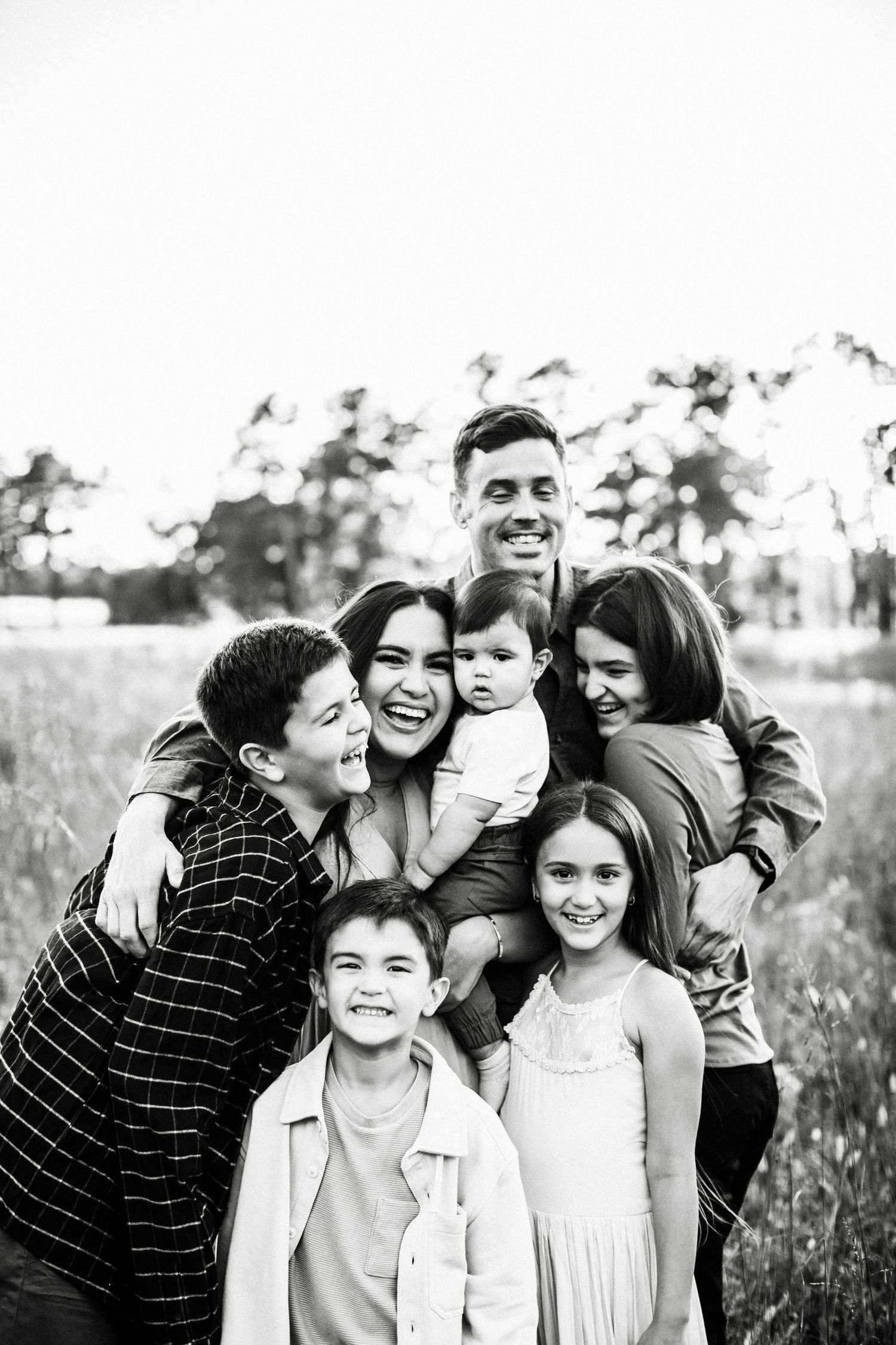 A joyful family poses together in a field with trees in the background, captured in black and white.