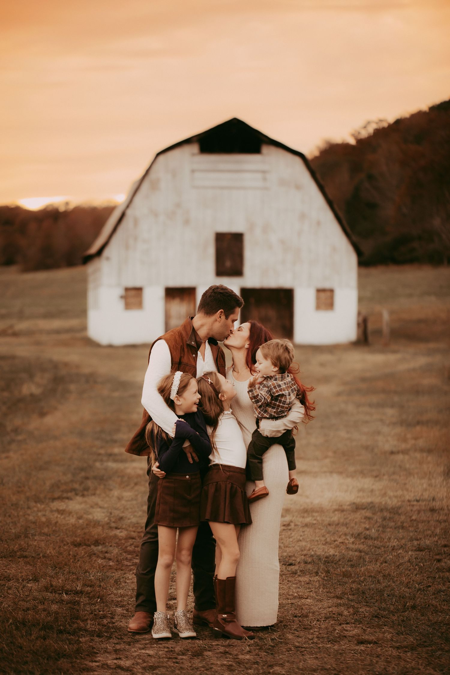 Family kissing at golden hour in front of a white barn on a rustic farm, with two young children nearby.