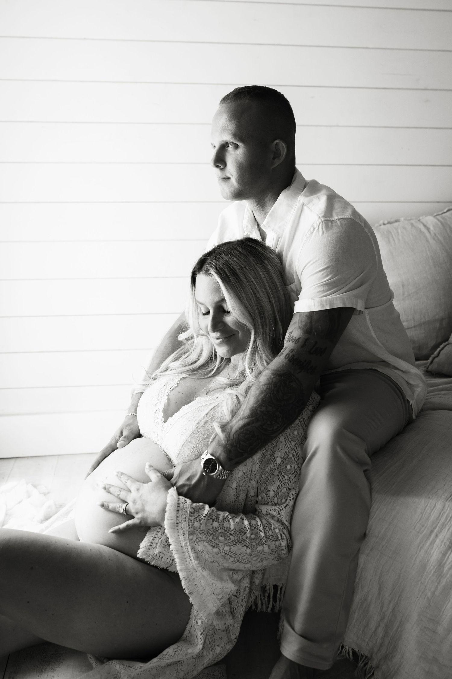 Black and white maternity photo of couple sitting together, man embracing pregnant woman from behind near white shiplap wall.