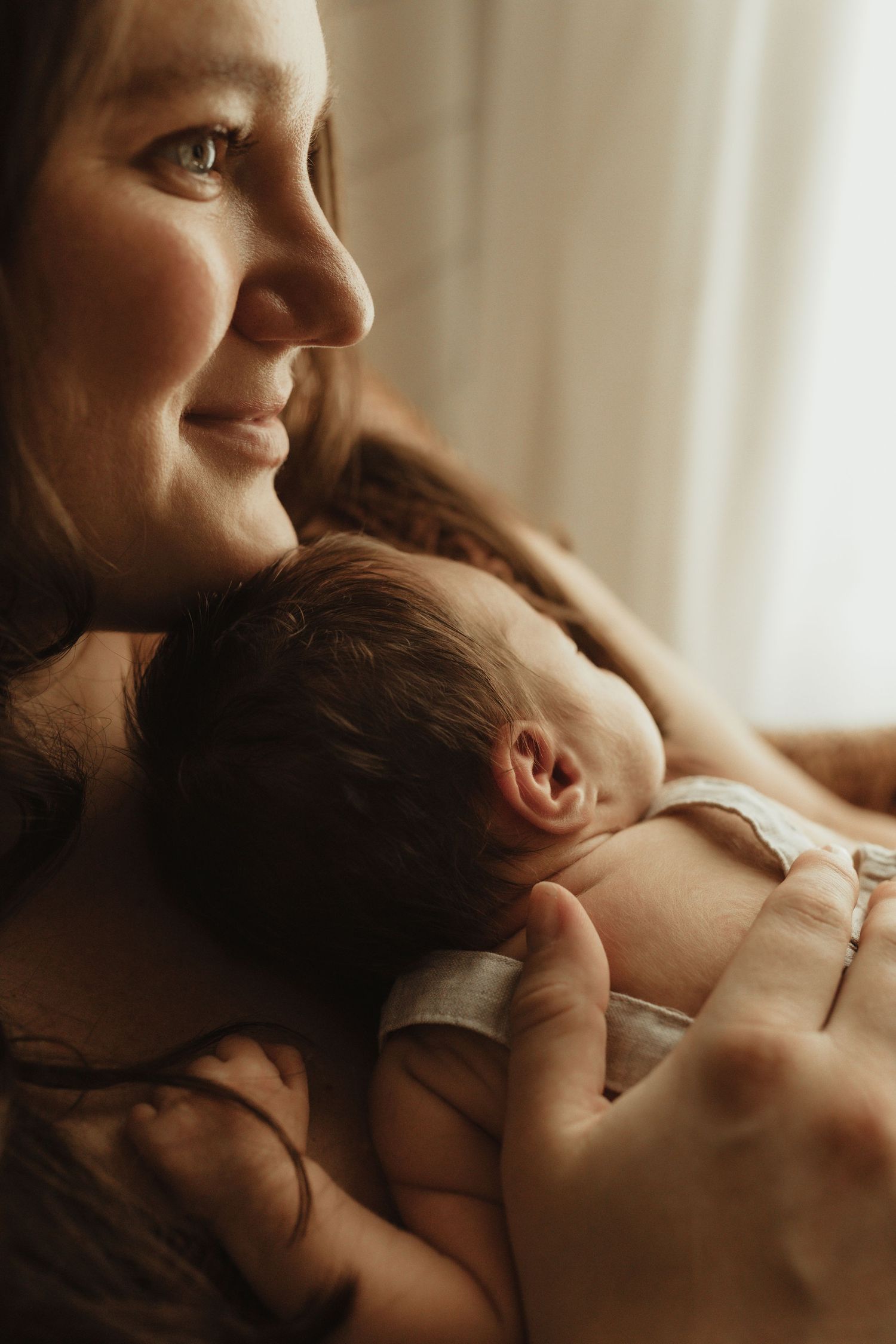 A smiling mother tenderly holds her sleeping newborn baby close to her chest in warm, soft natural light.