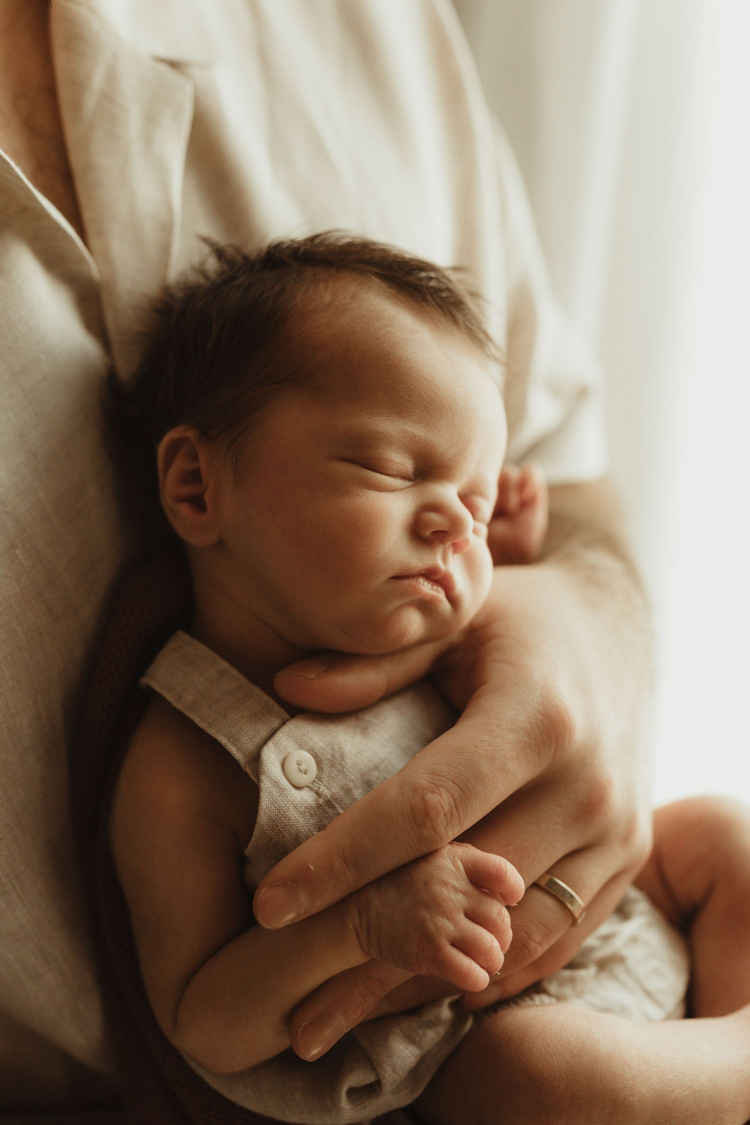 Newborn baby sleeping peacefully in parent's arms, wearing a gray romper, with soft natural light.