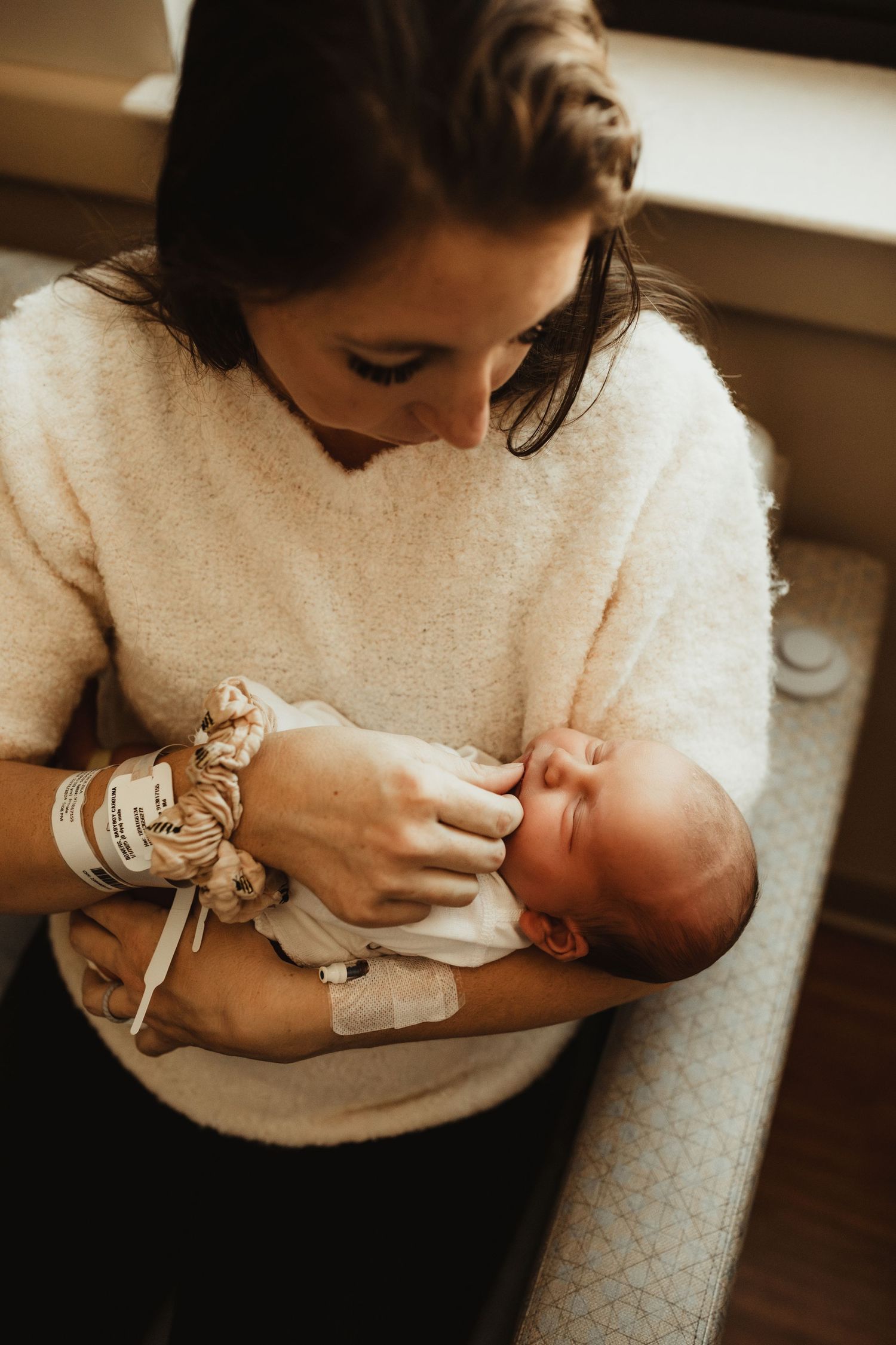 Parent holds and cuddles newborn baby in warm moody light while wearing a cozy white sweater.