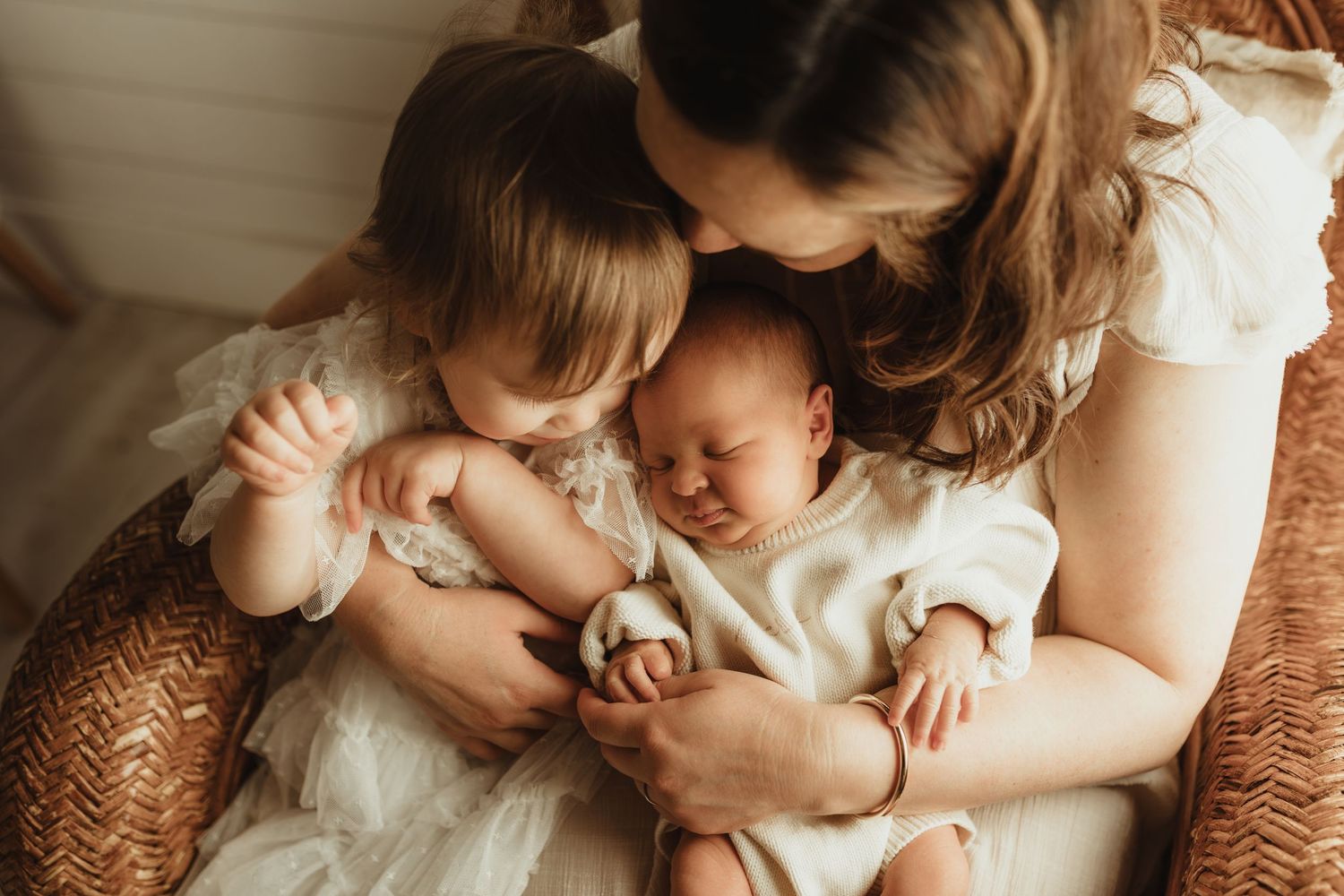 A mother tenderly holds her sleeping newborn while an older toddler in white dress leans in, family portrait.