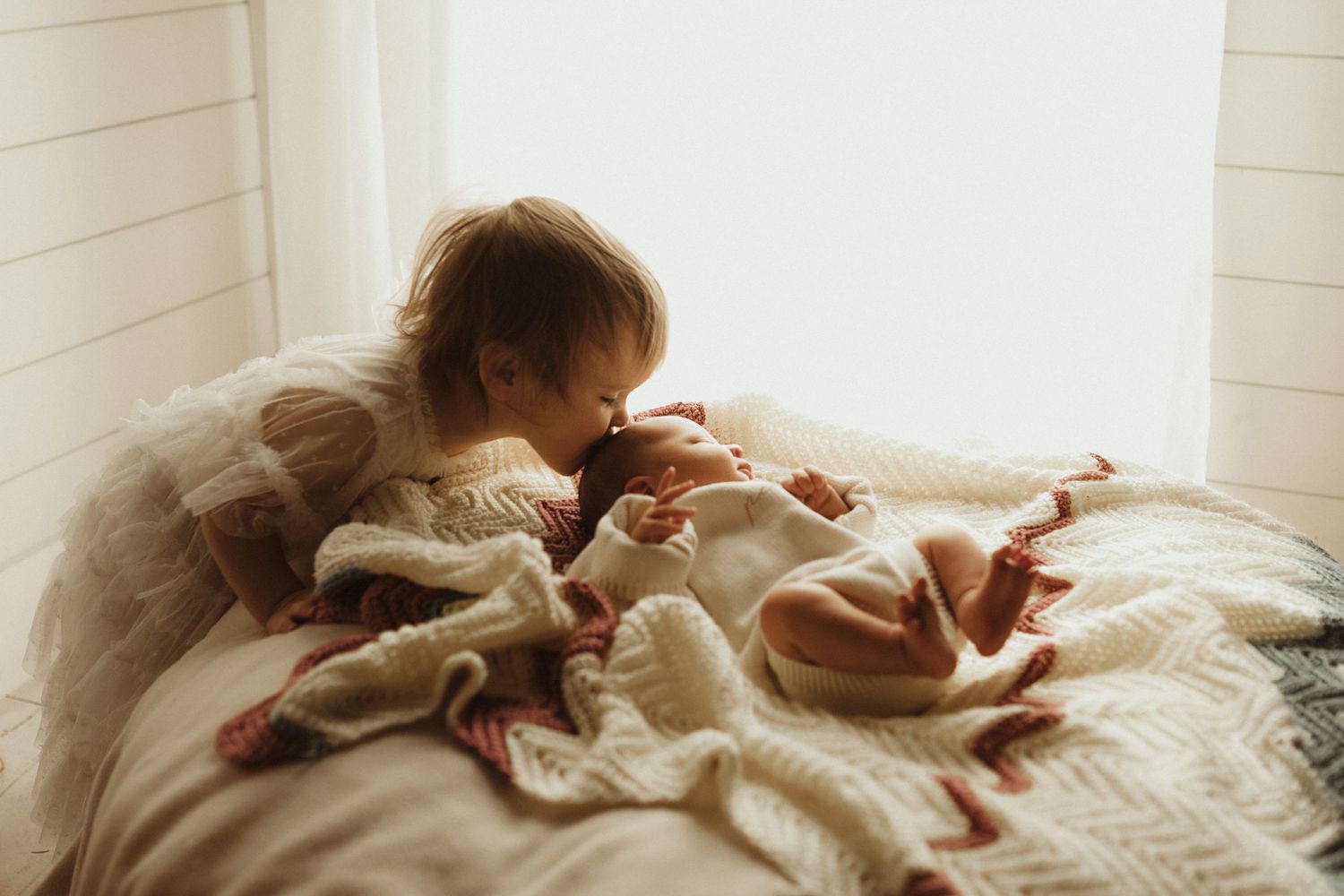 Young child in cozy sweater gently kissing newborn baby wrapped in cream knit blanket on a soft bed.