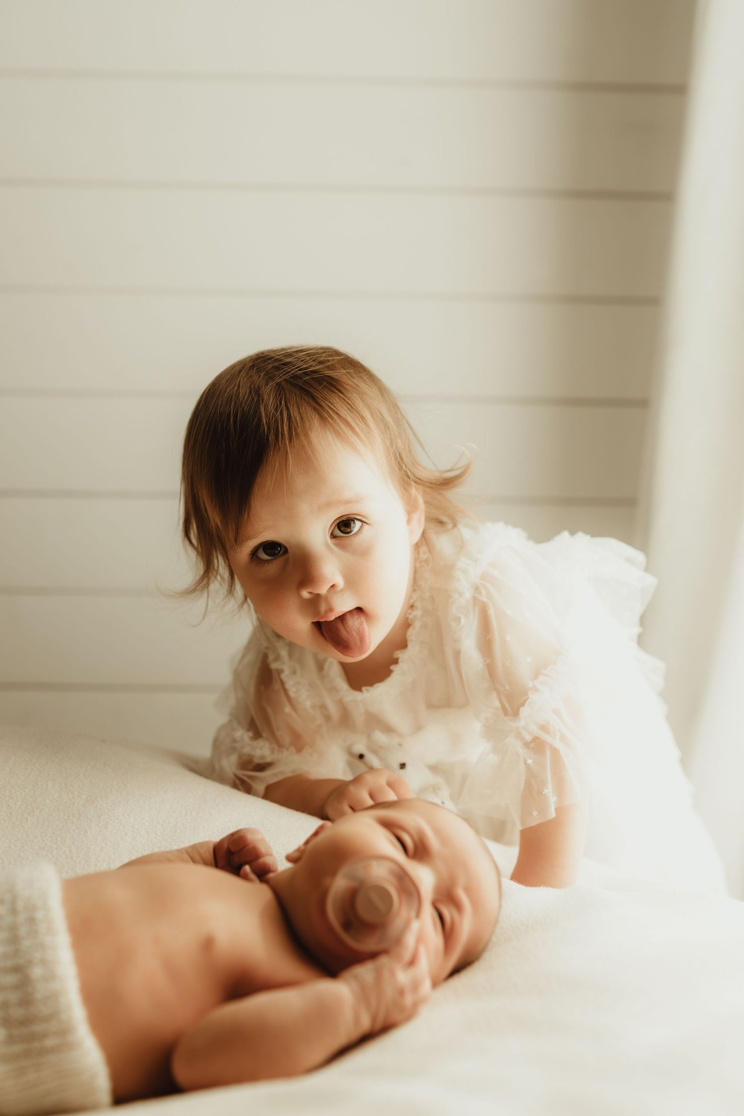 Toddler girl in white dress sticking tongue out while posing with sleeping newborn baby sibling on white bed.