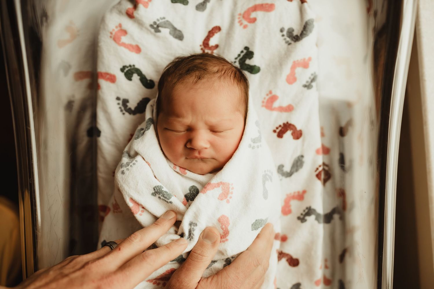 Newborn baby swaddled in a white blanket with colorful footprint pattern, sleeping peacefully in a bassinet.
