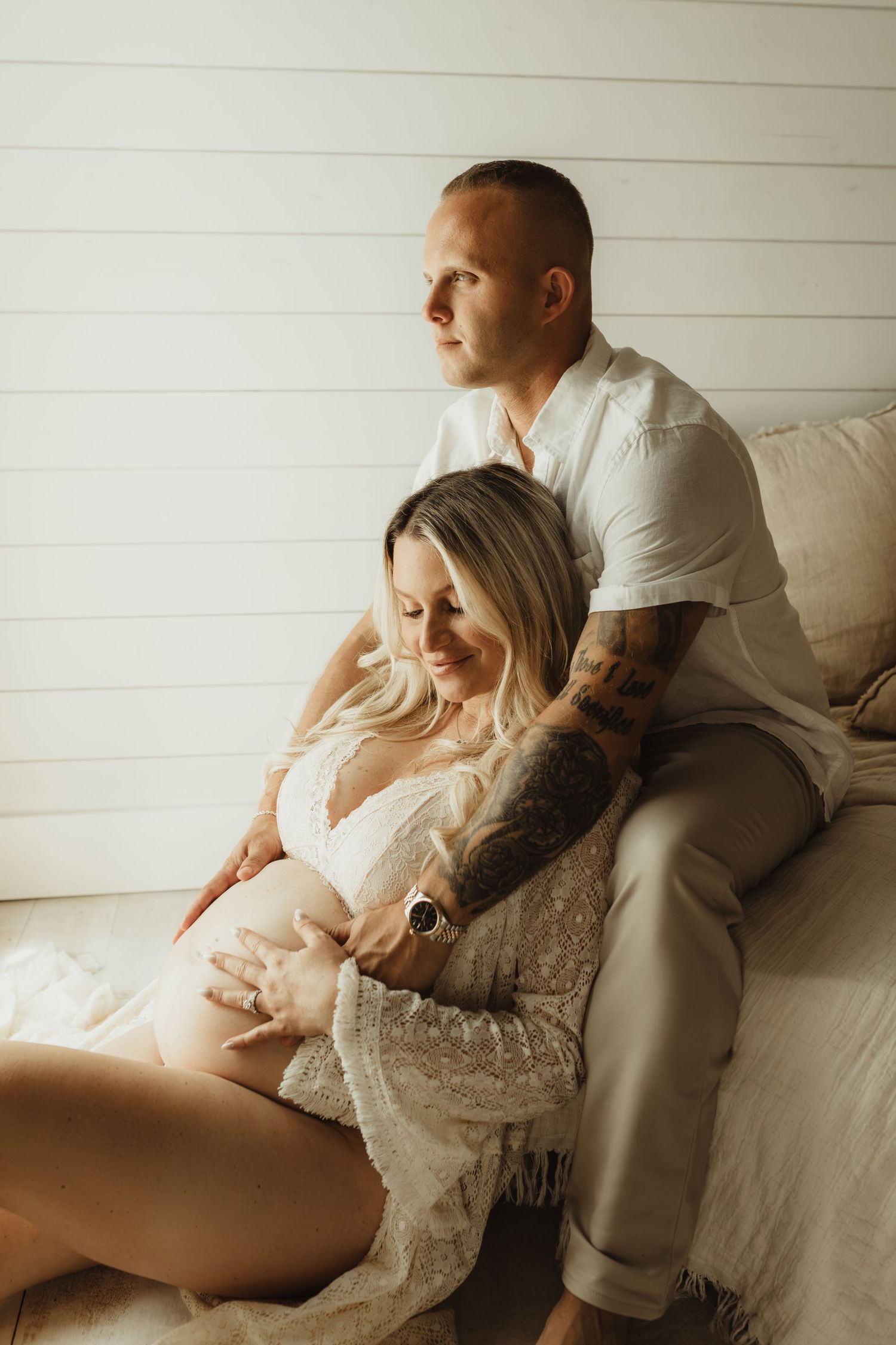 Couple sharing a tender maternity moment, tattooed man embracing pregnant woman in lace dress on cozy white backdrop.