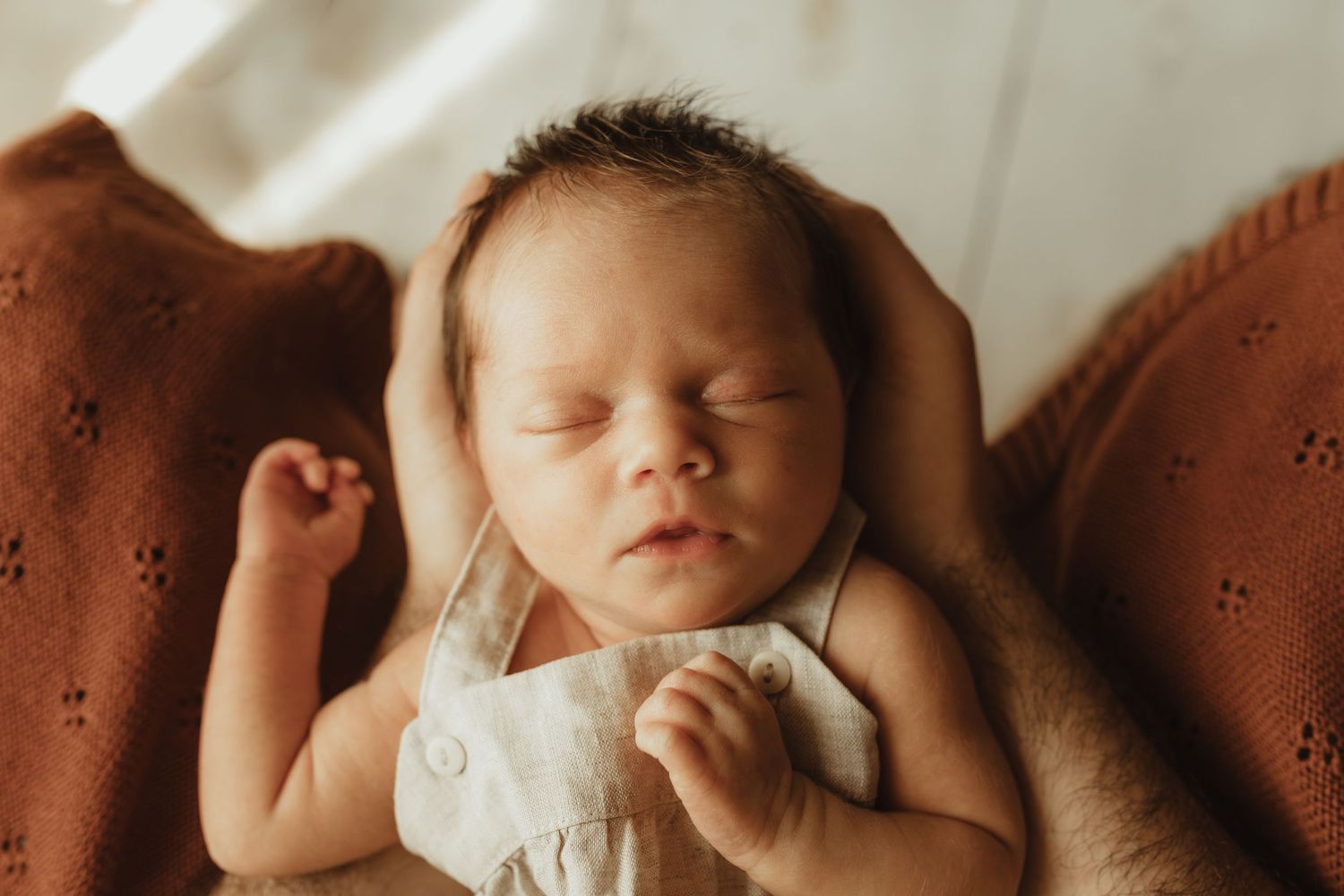 Peaceful newborn baby sleeping on a rust-colored knit blanket, cradled gently by adult hands.