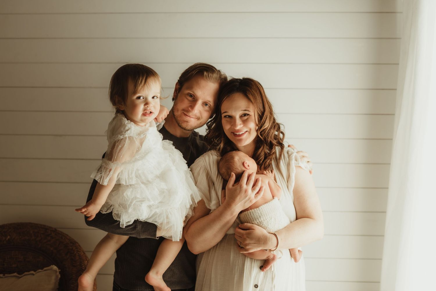 A young family of three poses warmly, with parents holding a baby girl in a white dress against a light shiplap wall.