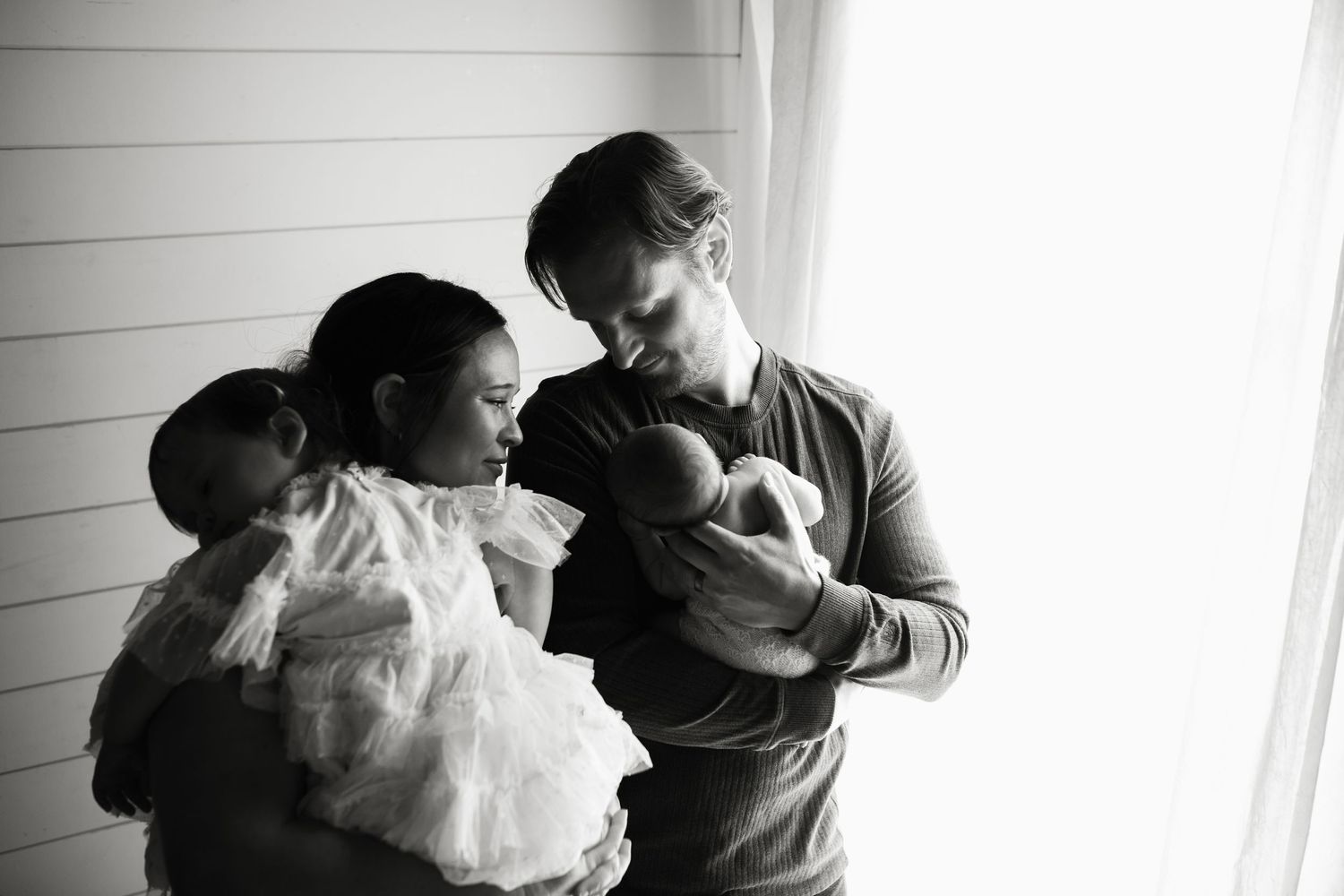 Black and white photo of parents holding newborn twins near a bright window.