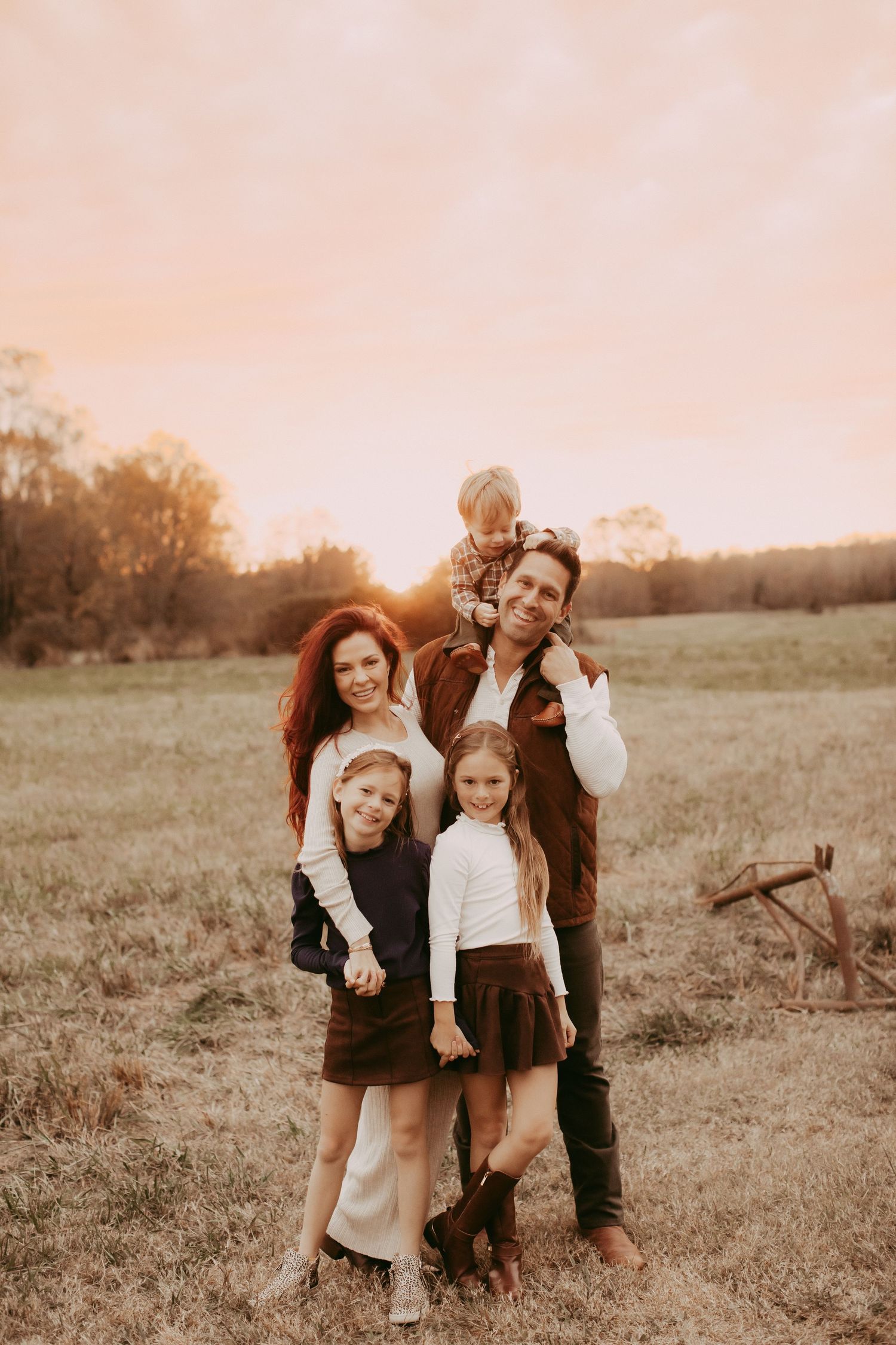 Family of five poses in golden sunset field, parents with three young children smiling outdoors in autumn.