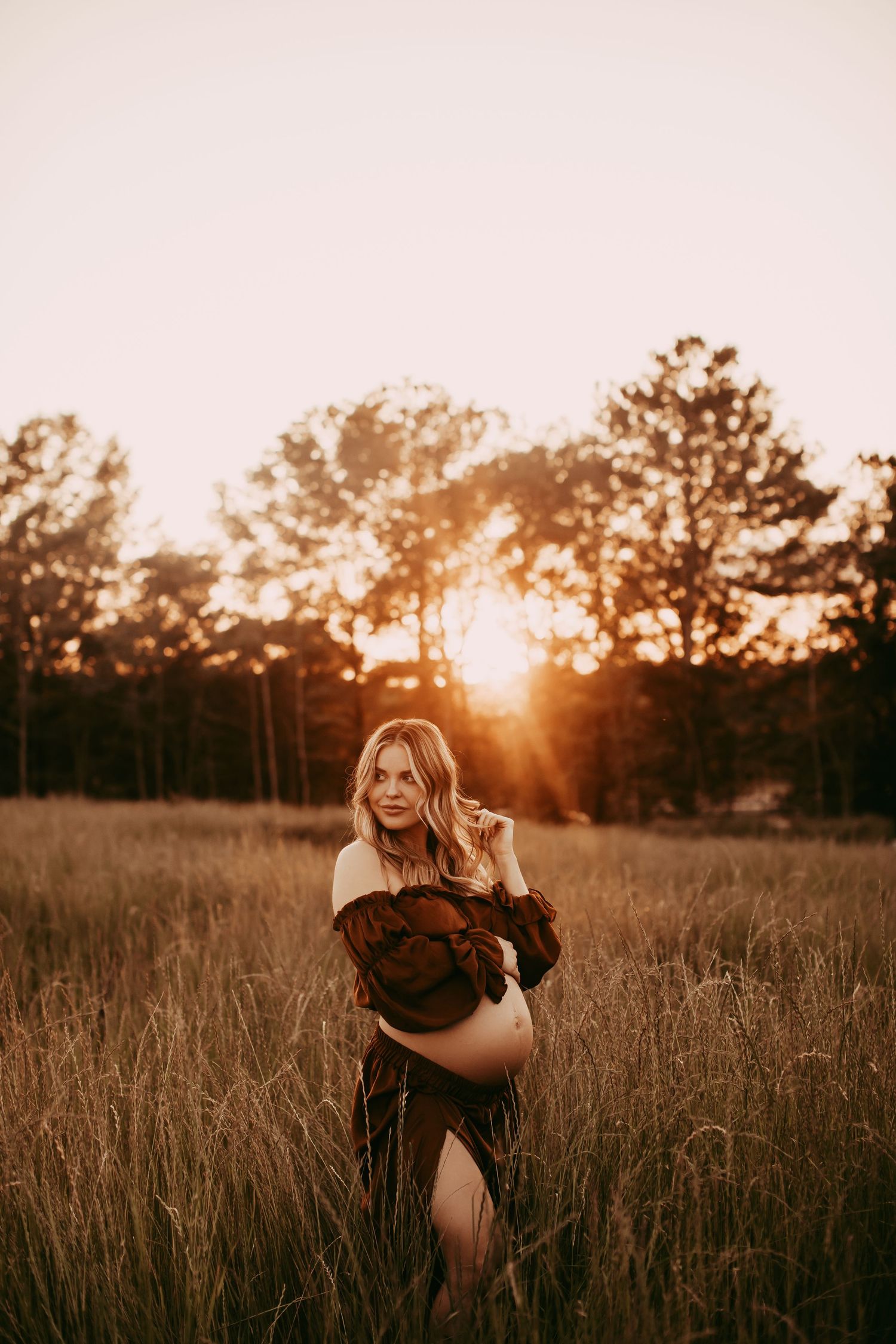 A silhouetted figure sits in a golden field at sunset with tall grasses and trees in the background.