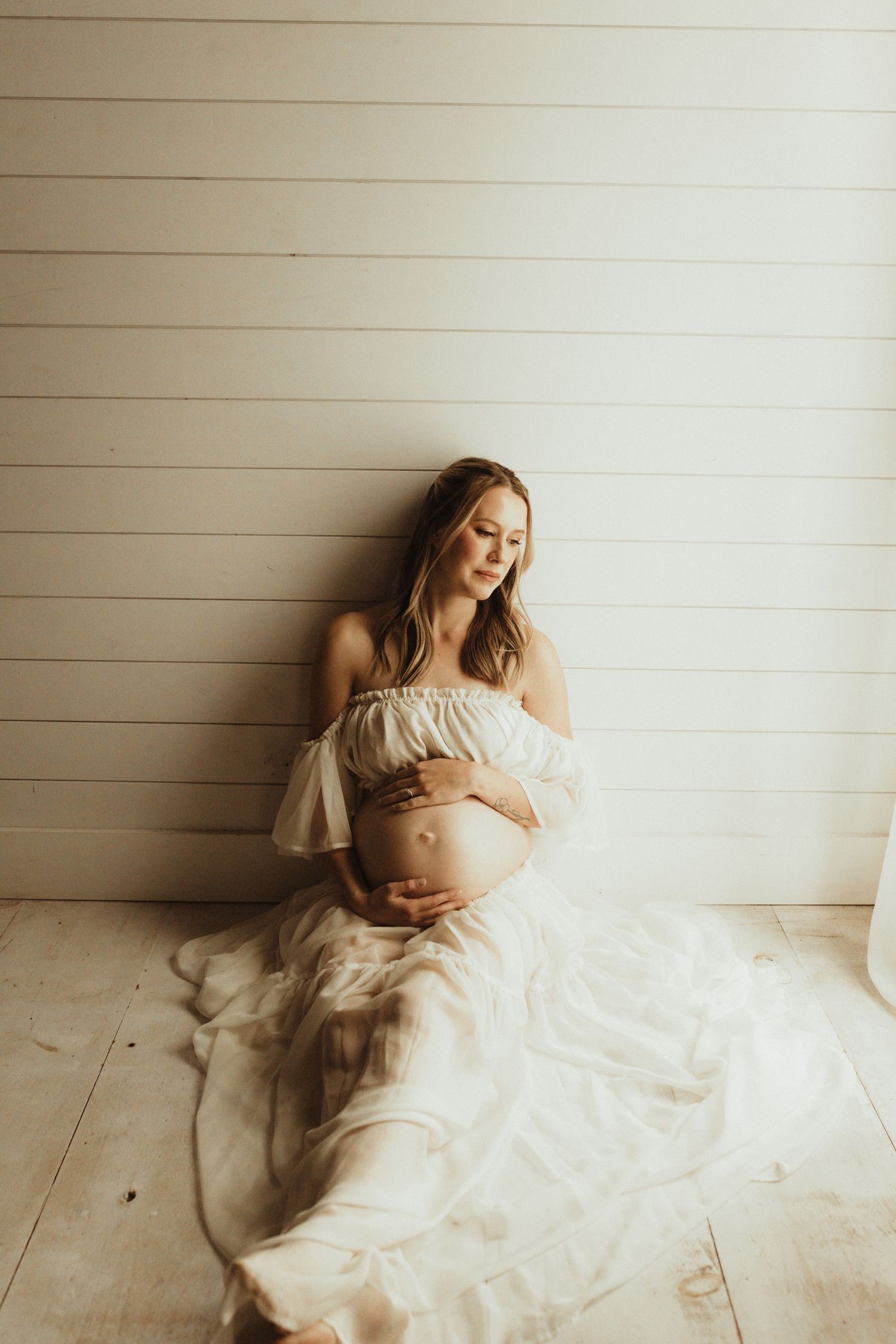 Artistic maternity portrait with ethereal white flowing dress against light wooden wall in natural lighting.