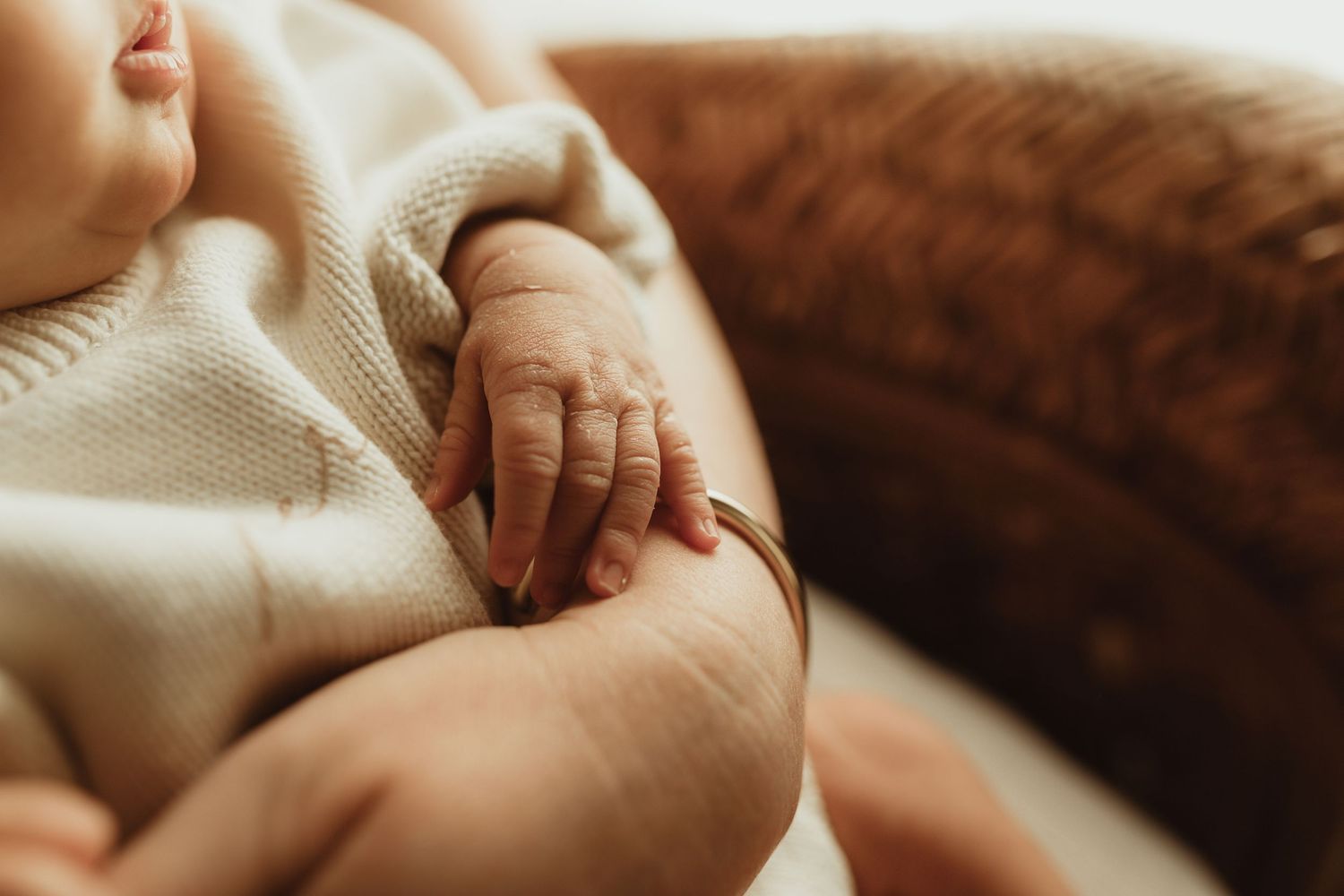 Close-up of a newborn baby's tiny hand gently resting on a parent's arm, wearing a cream knit outfit.