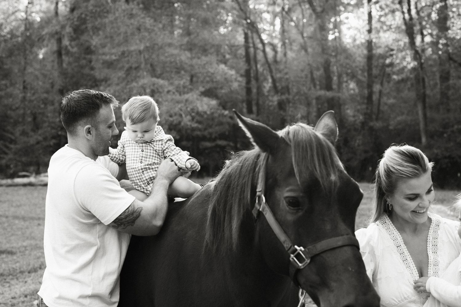 Black and white photo of a father placing a baby on a horse while a woman holds the reins outdoors.