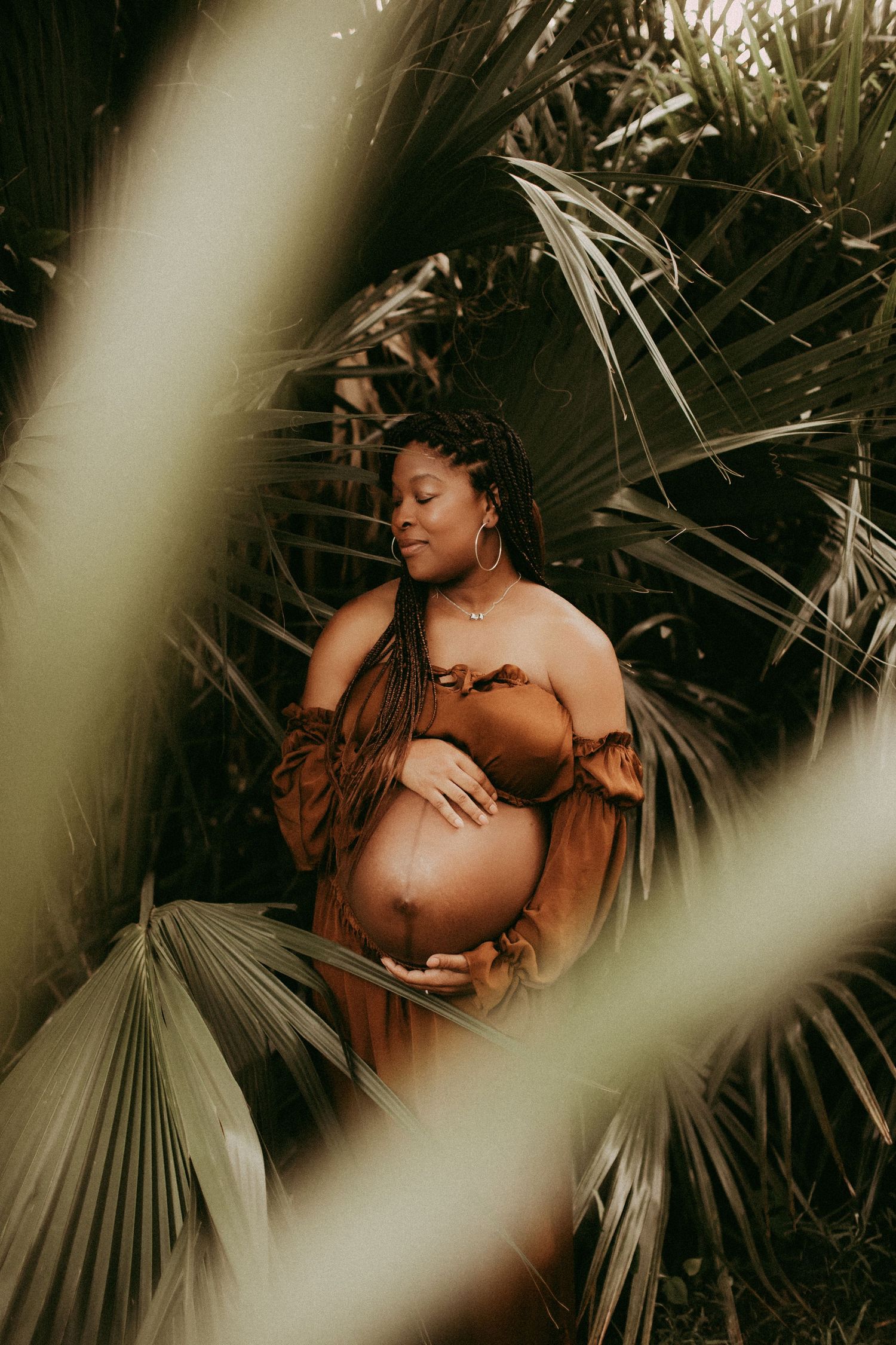Artistic maternity photoshoot in a tropical palm garden setting with warm brown tones and natural lighting.