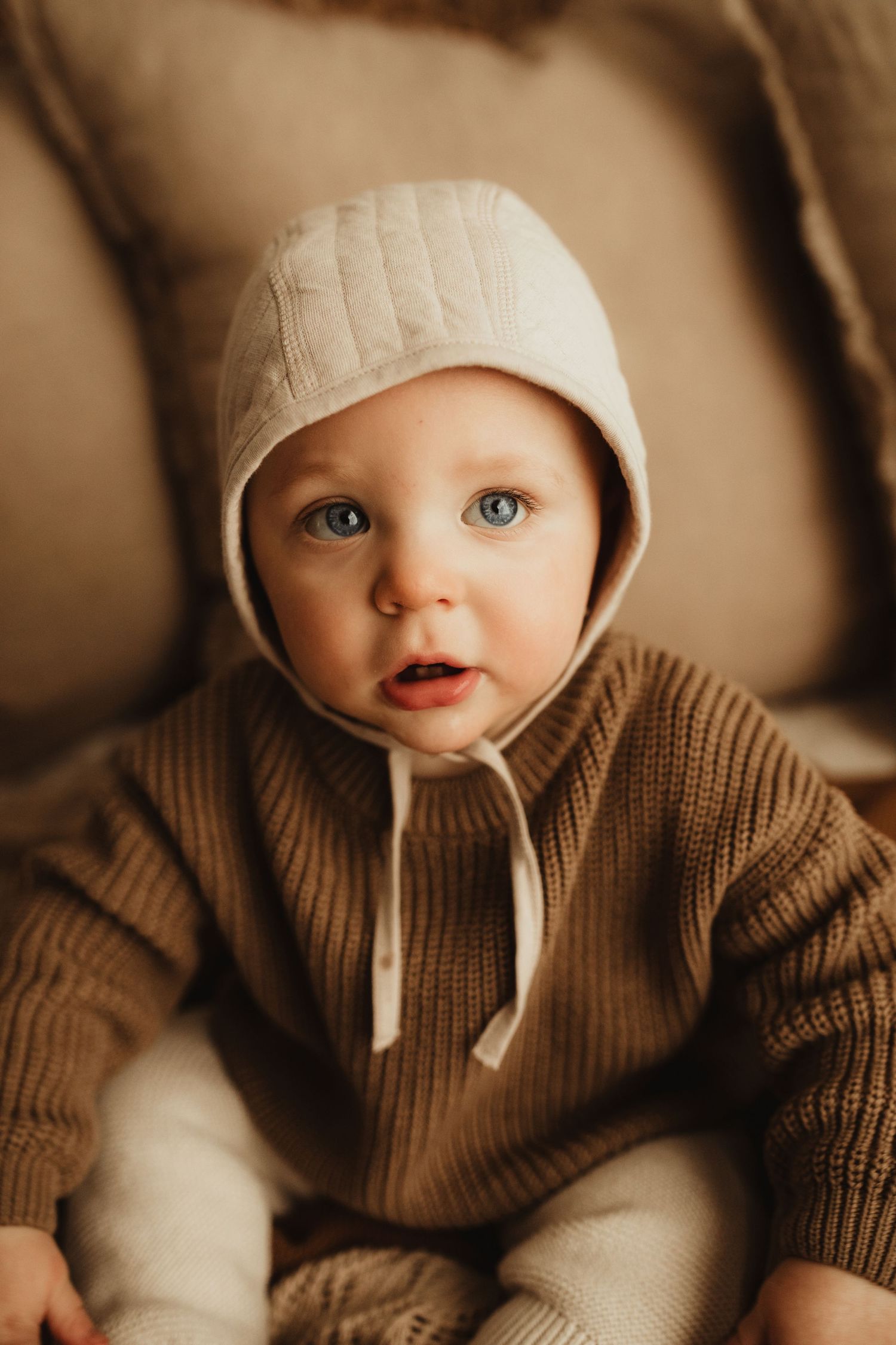 Baby wearing cozy brown knit sweater and beige beanie hat in warm sepia toned portrait series.
