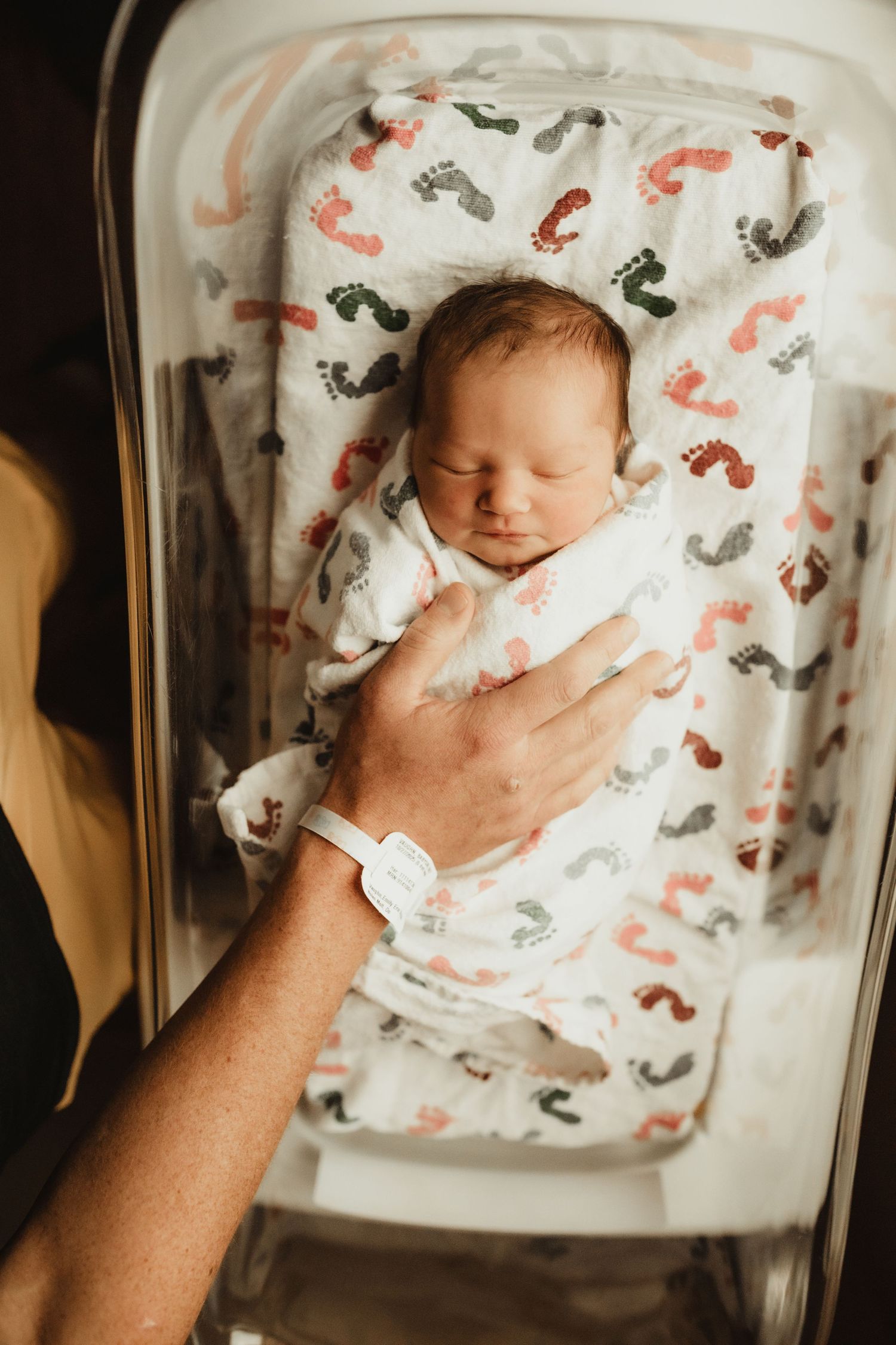 Newborn baby sleeping in hospital bassinet wrapped in footprint blanket with parent's hand resting gently on chest.