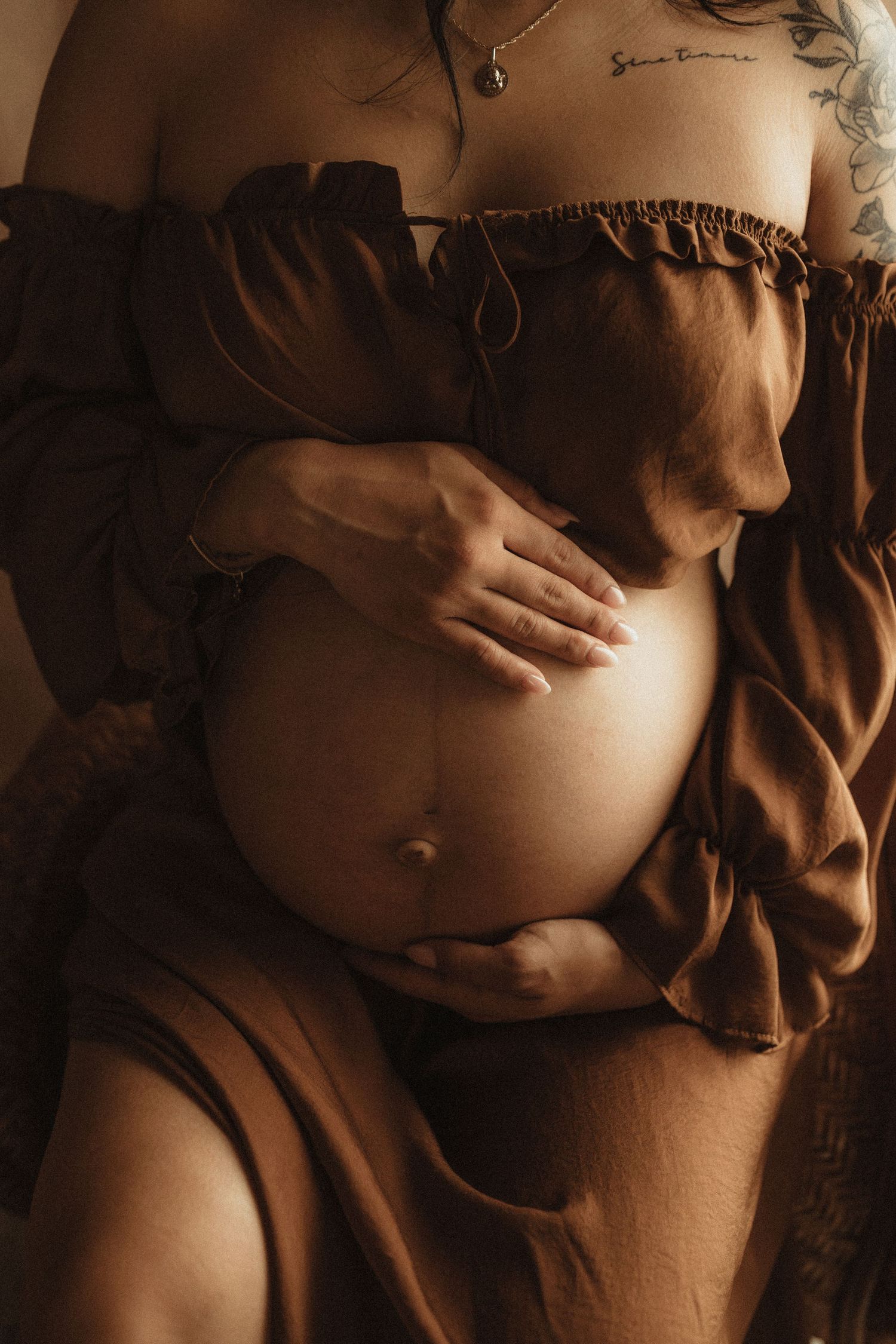 Pregnant woman in brown off-shoulder dress cradling her bare baby bump with both hands, warm toned maternity photo.