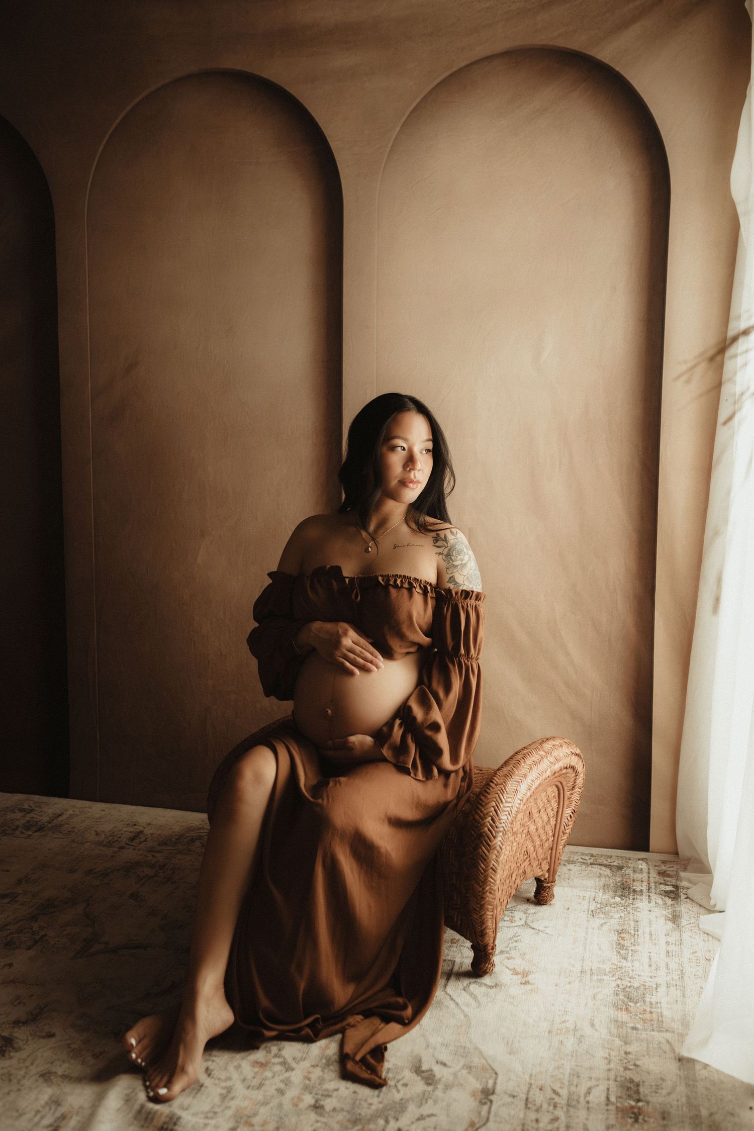 Pregnant woman in brown off-shoulder dress sitting gracefully in a warmly lit studio with arched backdrop.