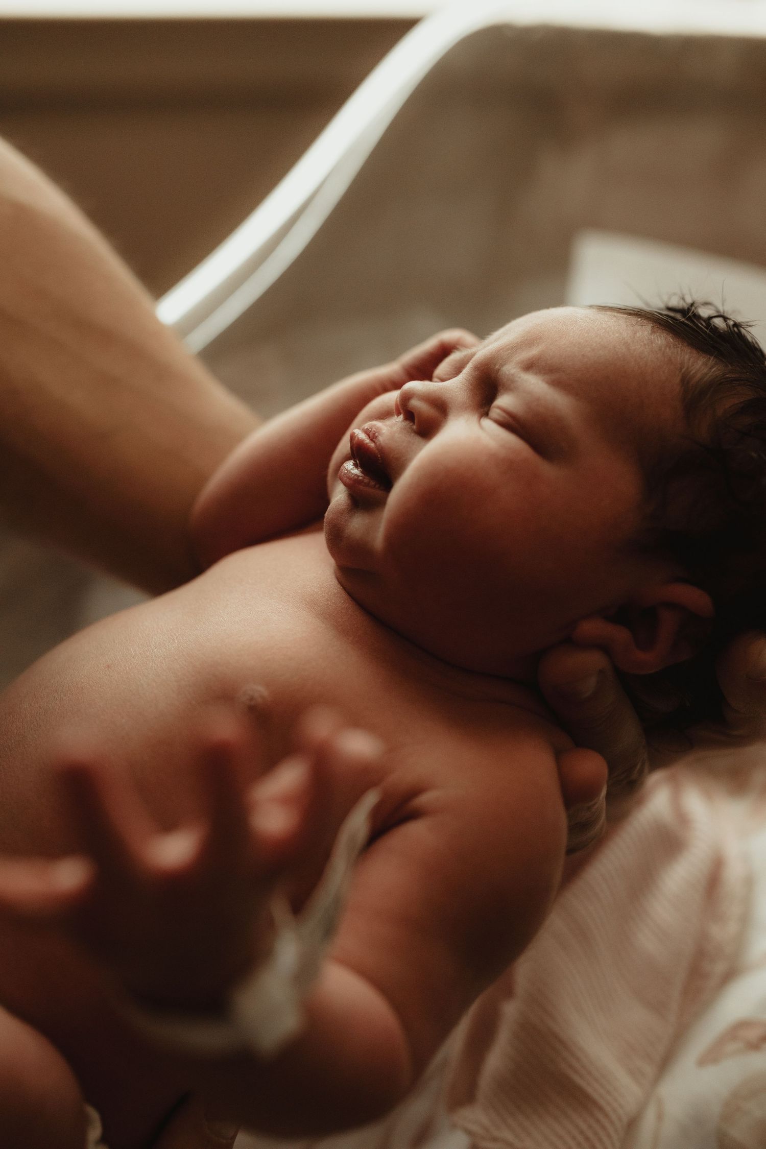 A newborn baby lying down with eyes closed while being gently touched by a caregiver's hand.